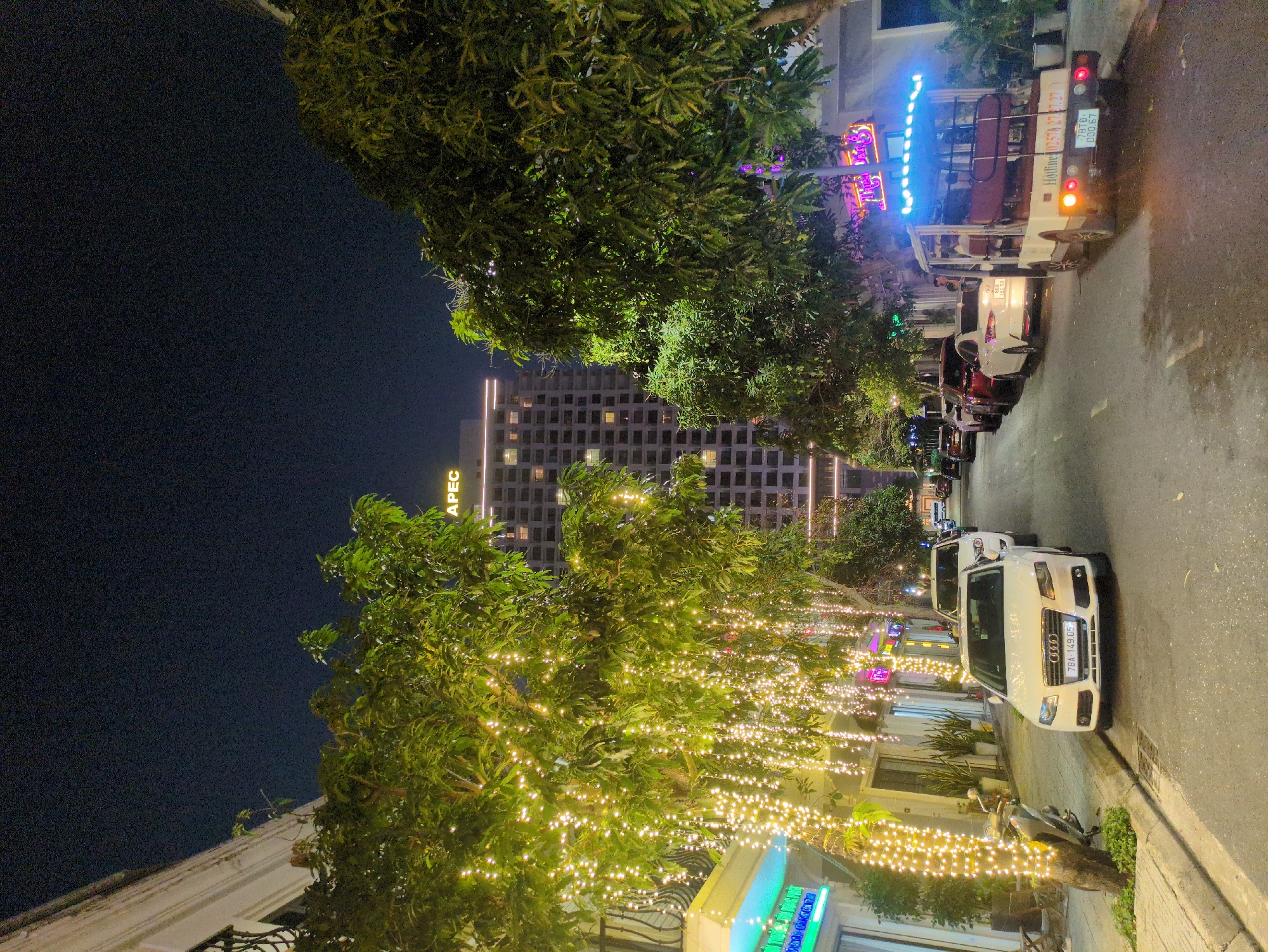 The image shows a street scene in Tuy Hòa, Vietnam, at night. The street is lined with buildings, trees, and parked cars. A tall building with the word "APEC" on the top is visible in the background. The trees are decorated with strings of lights. A white Audi car is parked on the side of the road with the number plate 78A-149.05. A motorized rickshaw with a decorative light display is on the street, and a row of parked vehicles can be seen further down the road.