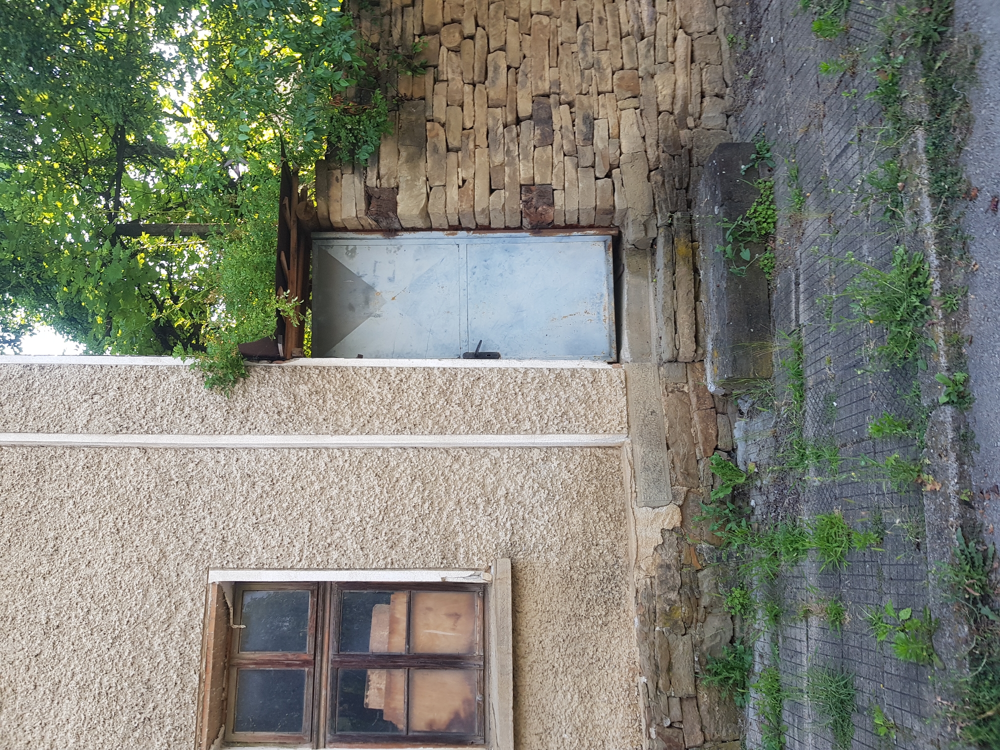An exterior shot captures a weathered building facade in Lomets, Bulgaria. A single, light-blue metal door is set into the building, flanked by a rough-hewn stone wall on the right and a textured, pale stucco wall on the left. The door has a black handle and appears to be slightly ajar, with a small, weathered metal overhang above it. To the left of the door, a multi-paned wooden window with dirty glass offers a glimpse of objects stacked inside. The building's base is constructed with rough stones, and the ground in front is a gray, grid-patterned paved area interspersed with weeds and grass. A large, dark gray concrete block sits to the right of the doorway. Overhead, the dense green foliage of trees partially obscures the sky.