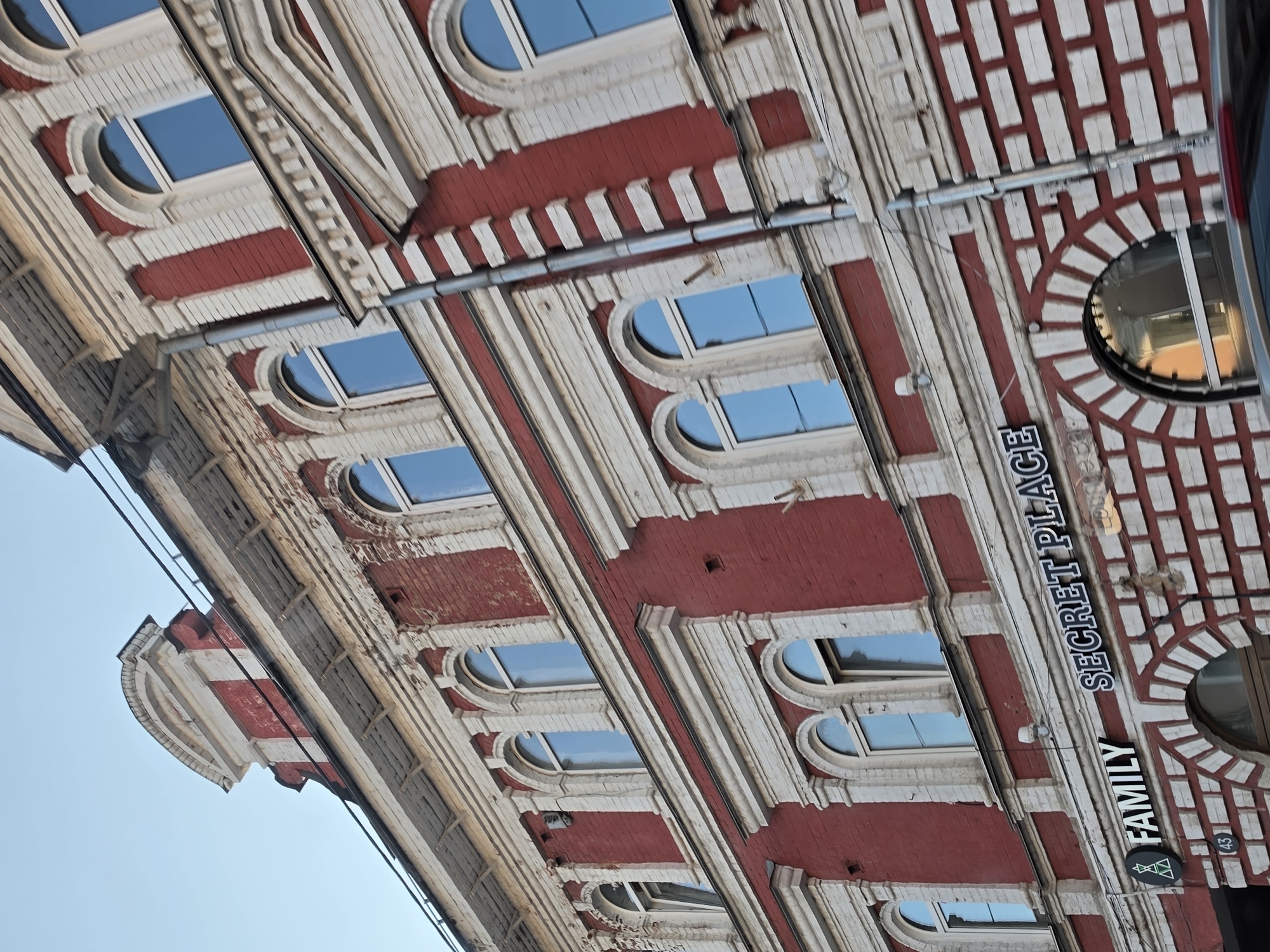 This is a low-angle, exterior, medium shot of the facade of a historic brick building in Nizhny Novgorod, Russia. The building features red brick walls with white decorative architectural elements, including arched window frames and horizontal bands. Several arched windows with blue glass are visible across the facade, some of which appear to be modern replacements. On the ground level, signage is visible. To the left, a sign reads "FAMILY" with a stylized green logo. To the right of that, a sign reads "SECRET PLACE." The sky above is a clear, light blue. The image is slightly tilted, as if taken from a moving vehicle or with a handheld camera. The overall impression is of an old, but maintained, urban building.