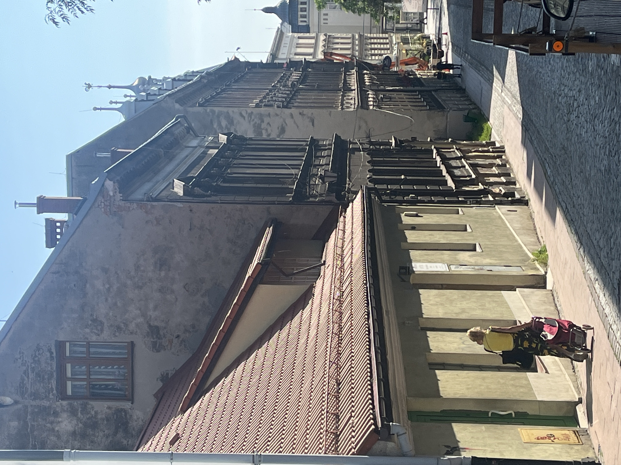 This is a vertically oriented photograph of an urban street scene. The foreground shows the roof of a building with red tiles and a large skylight. To the left of this roof is the facade of a grey building with a small, multi-paned window. Further into the background, a large, ornate building with intricate architectural details is visible. Construction equipment, including an orange excavator, is present in front of this building, suggesting ongoing renovation or demolition work. Beyond this, a row of buildings with classical facades lines the street.
In the middle ground, a woman is pushing a shopping cart along a paved walkway. She is wearing a yellow shirt and a patterned skirt. The sun is casting long shadows, indicating that it is likely late afternoon. The sky is clear and blue, suggesting fair weather. The overall scene conveys a sense of everyday life unfolding in a historic European city, with elements of construction adding a dynamic aspect to the urban landscape. There is no visible text in the image.