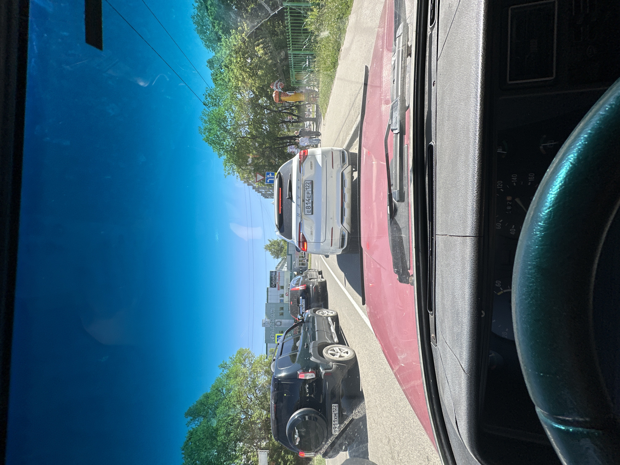 This image, taken from inside a vehicle in Novoaltaysk, Russia, captures a daytime scene of a traffic jam. The perspective is from the driver's seat, with the steering wheel visible in the foreground. Through the windshield, several cars are lined up on a road, including a white SUV with the license plate E814EP 122 and a dark SUV with the license plate 8911XM 122. In the background, there are trees and buildings, one of which has the text "ΦΛΑΓΜΑΗ" and "Garderob" visible. There are also some people walking on the sidewalk, and inflatable figures, possibly for decoration or advertising, are present. The sky is a clear blue, suggesting fair weather. The overall atmosphere appears to be calm, despite the traffic.