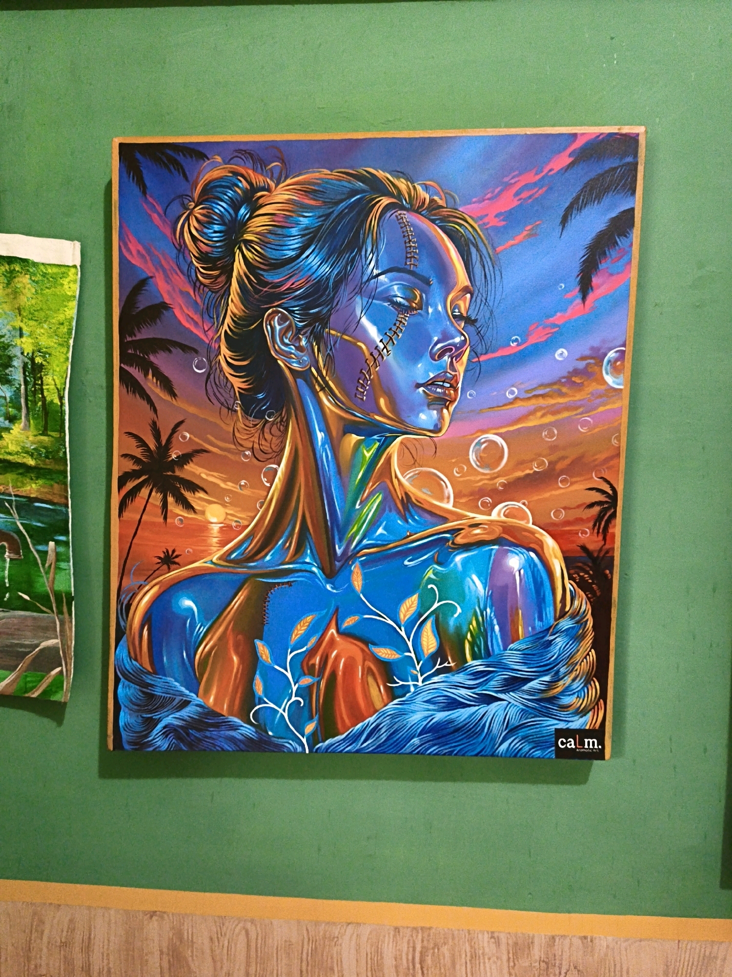 In Pabelan, Indonesia, a framed painting hangs on a textured green wall. The artwork depicts a woman with her eyes closed, facing right. Her skin is a vibrant blend of blues, oranges, and purples, reflecting a dramatic sunset sky filled with pink and orange clouds. Palm trees silhouette against the sky, and several translucent bubbles float in the air. The woman's hair is pulled up in a bun, and she wears a metallic earring. Her face has visible stitching detail, and there are lighter-colored vine-like growths on her body. The lower part of the painting shows her shoulders covered by what appears to be swirling water or fabric. A small black rectangular label with the word "cal.m." is affixed to the bottom right corner of the painting. To the left of this painting, a portion of another artwork is visible, featuring a green landscape with trees and a body of water. The wall is bordered at the bottom by a light brown, wood-grain pattern.