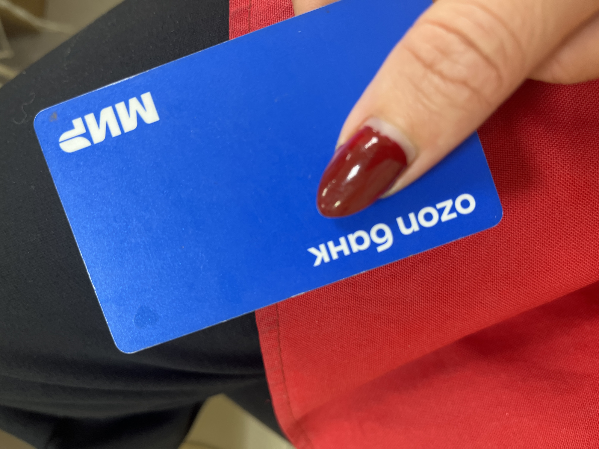 The image shows a close-up of a person's hand holding a blue card with white text. The card has the "Мир" logo on the top right and "ozon банк" written vertically along the left side. The person's fingernail, painted a dark red, is visible near the center of the card. The card is positioned over a red fabric surface, possibly an apron or garment, which is in turn against a dark fabric background, likely clothing. The overall scene is an indoor, personal setting, with no discernible elements placing it specifically in Blagoveshchensk, Russia, or any other city. The lighting suggests an artificial indoor environment.
