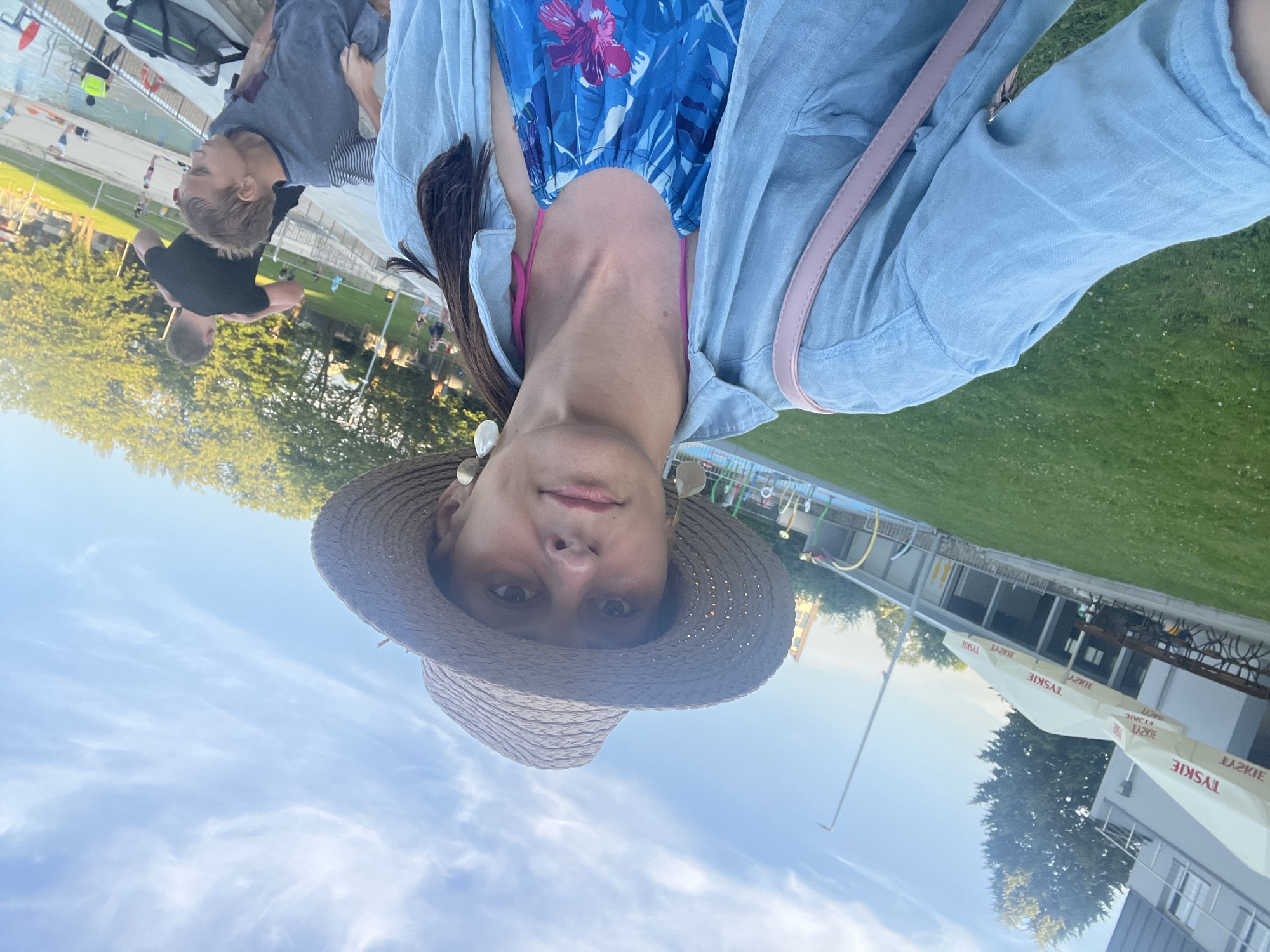 An outdoor selfie is taken by a woman wearing a wide-brimmed straw hat, large circular earrings, a light blue button-down shirt, and a blue and pink floral patterned halter top. She is standing in front of a grassy area and a body of water, possibly a pool or water park. In the background, there are trees, a building with umbrellas displaying "TYSKIE," and several other people, including a man in a black t-shirt and a young boy in a striped shirt. The sky is blue with wispy clouds, and the sun appears to be setting, casting a warm glow on the scene. The woman is holding a pink purse with a strap across her chest.