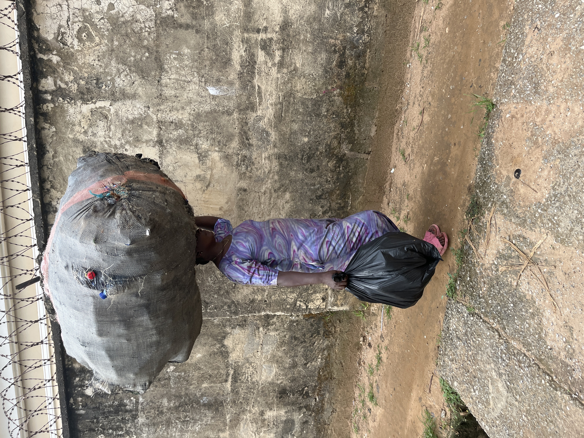 A woman carries a large, heavily filled sack of what appears to be fabric or refuse on her head, while holding a black plastic bag in her right hand. She is walking on a dirt path next to a concrete structure and a weathered, grey concrete wall. A barbed wire fence is visible along the top of the wall. The woman is wearing a colorful, patterned dress and pink sandals. The environment suggests an urban or semi-urban setting, possibly in a less developed area of Abuja, Nigeria, given the construction materials and the presence of the wall and fence. The overall scene depicts a person engaged in carrying heavy loads, a common sight in many parts of the city.