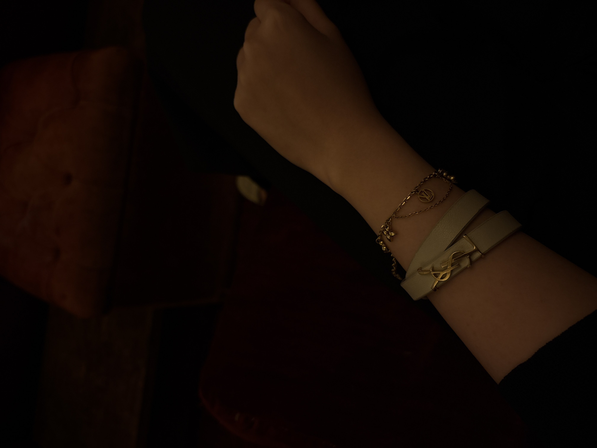 A right arm and hand are visible in the image, situated in a dimly lit environment. The arm is adorned with two bracelets. One is a delicate gold chain with a pendant that looks like the Louis Vuitton logo. The other bracelet appears to be a light-colored leather wrap bracelet with a gold Yves Saint Laurent logo. The arm is positioned against a dark background, which has some vague hints of furniture on the side, as if a part of a chair. The location is unknown.