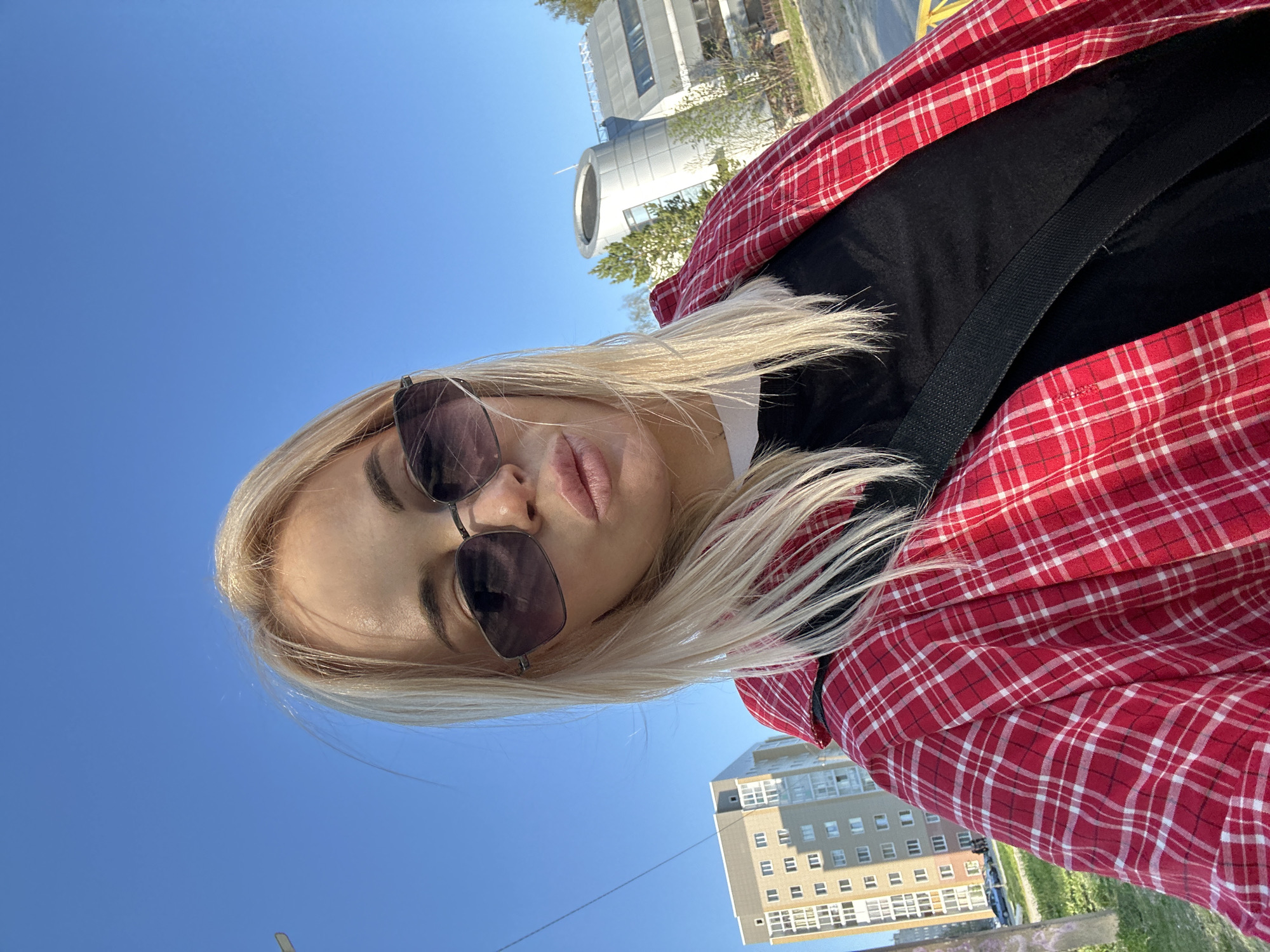 A young woman with blonde hair is posing for a selfie outdoors. She is wearing sunglasses and a red and white checkered flannel shirt over a black top. A black strap crosses her chest, possibly a purse or bag strap. The background shows a clear blue sky and several buildings, including a cylindrical structure with a white roof and a gray building with numerous windows. Based on the buildings and general environment, the location is likely in the city of Megion, Russia.