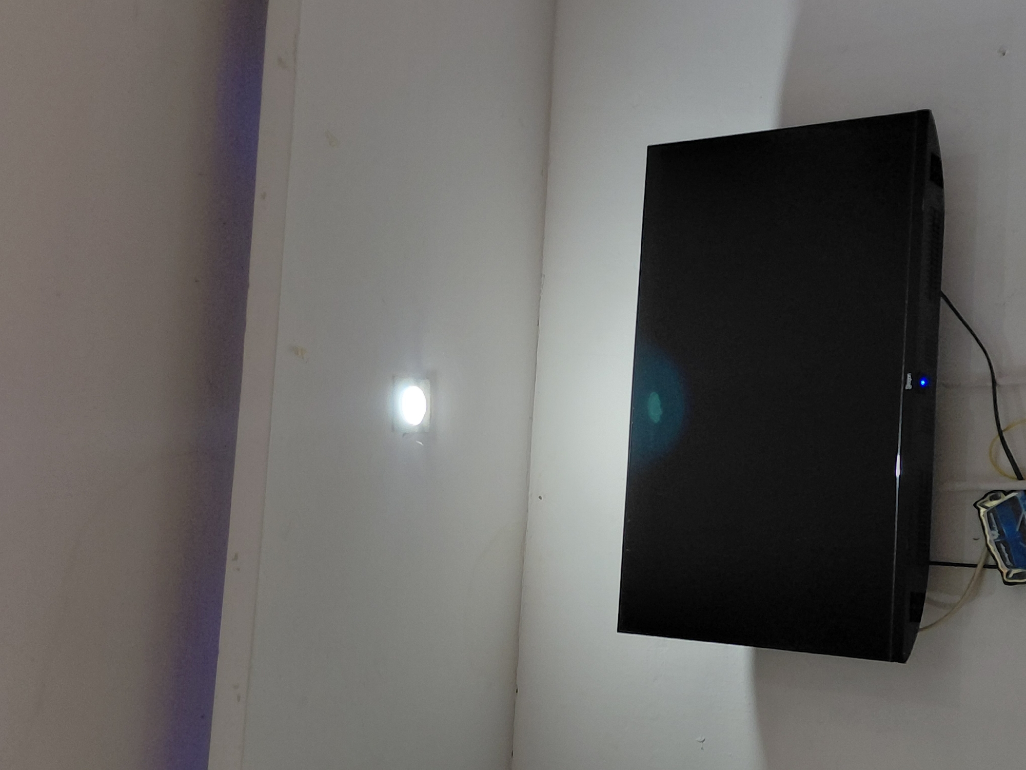 The image captures an indoor corner where two white walls meet, illuminated by various light sources. On the right wall, a rectangular, black electronic device, resembling a monitor or television, is mounted. A small, bright blue indicator light is visible on its right side. Several wires, some black and some yellow, extend from the back or side of this device, trailing downwards and connecting to a white, blue, and black electrical fixture or junction box with a somewhat decorative or protective cover.
On the left wall, a circular, recessed light fixture emits a very bright white light, creating a strong hotspot on the wall. Further to the left, in the corner where another wall meets the main left wall, a vertical strip of light casts a subtle purple glow, suggesting additional ambient lighting. The walls appear to be painted white, with some minor imperfections or stains visible.
No people are present in the image, and no specific activity is taking place. The scene appears to be a stationary setup of electronic equipment and lighting within a room. The overall lighting suggests an indoor setting where artificial light sources are primary. There is some faint, illegible text or a logo visible on the bottom right edge of the black electronic device. The location is an indoor space in Santa Ana, Venezuela.