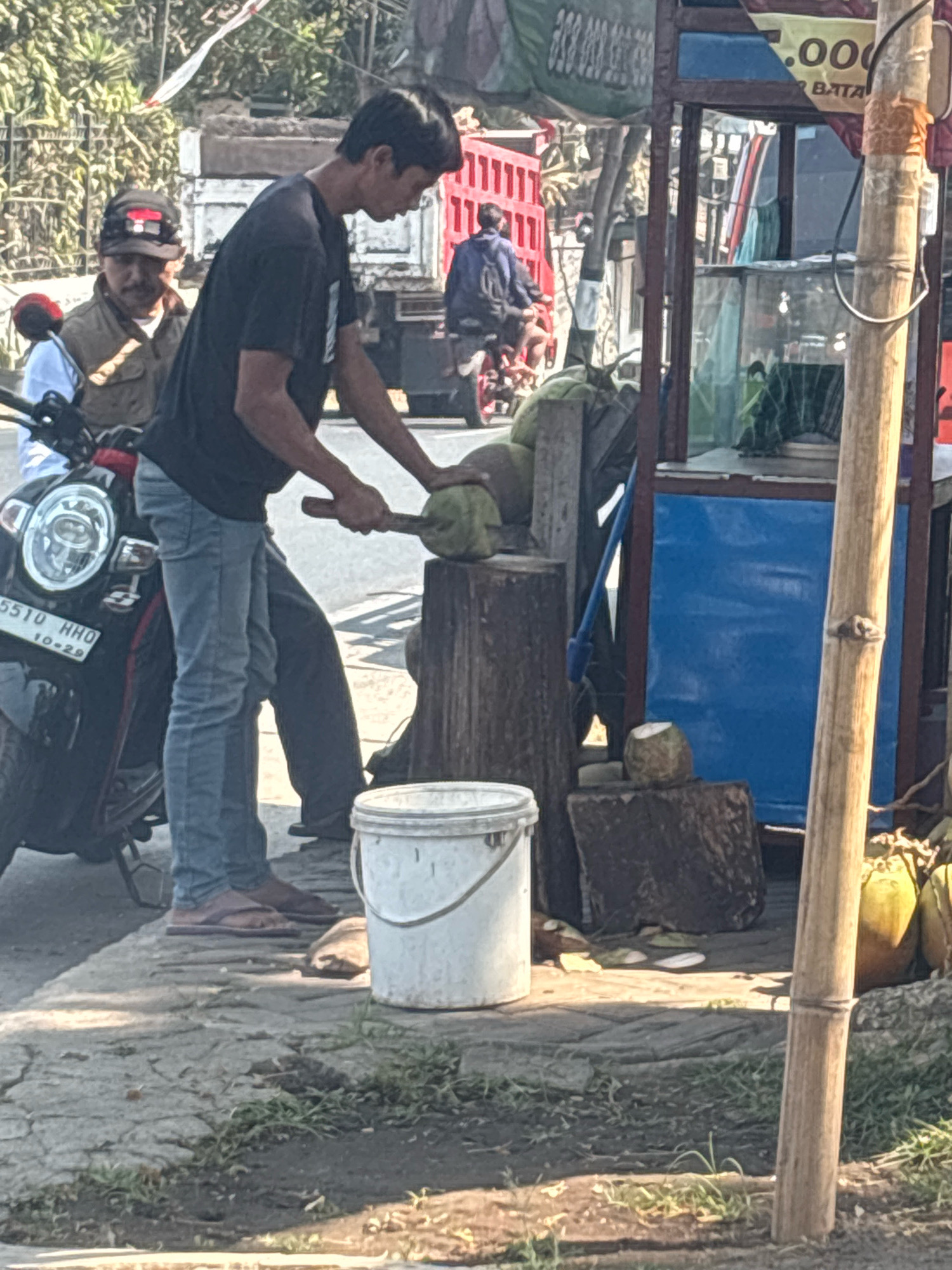 In Belung, Indonesia, a young man in a black t-shirt and blue jeans is standing by a scooter, preparing to cut a coconut. He holds a long, thin knife in his right hand, poised above a green coconut that is secured in a wooden stump. Next to the stump is a white plastic bucket and several more coconuts. In the background, a man wearing a brown jacket and a cap is seated on the scooter. Behind them, a blue and red food stall is visible, with a sign that has "000" printed on it. The ground is paved with rough concrete, and patches of grass are growing between the stones. The overall scene depicts a typical street-side vendor preparing a refreshing drink.