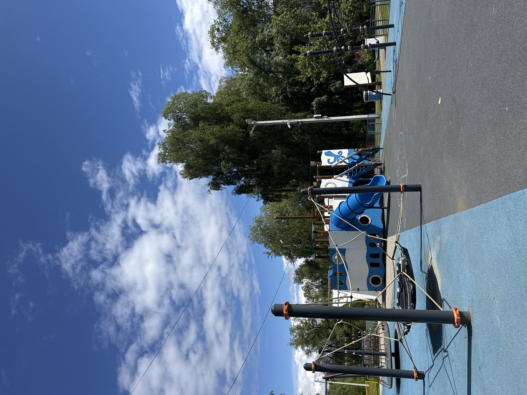 The image shows a playground in Shevlyakovo, Russia. The playground has a blue and white play structure resembling a ship with a blue slide. Rope structures are connected to black poles with orange caps. The ground is a combination of blue and gray rubberized material. A line of tall trees is visible in the background, and a partly cloudy blue sky is above. A pole with electrical equipment is also visible.