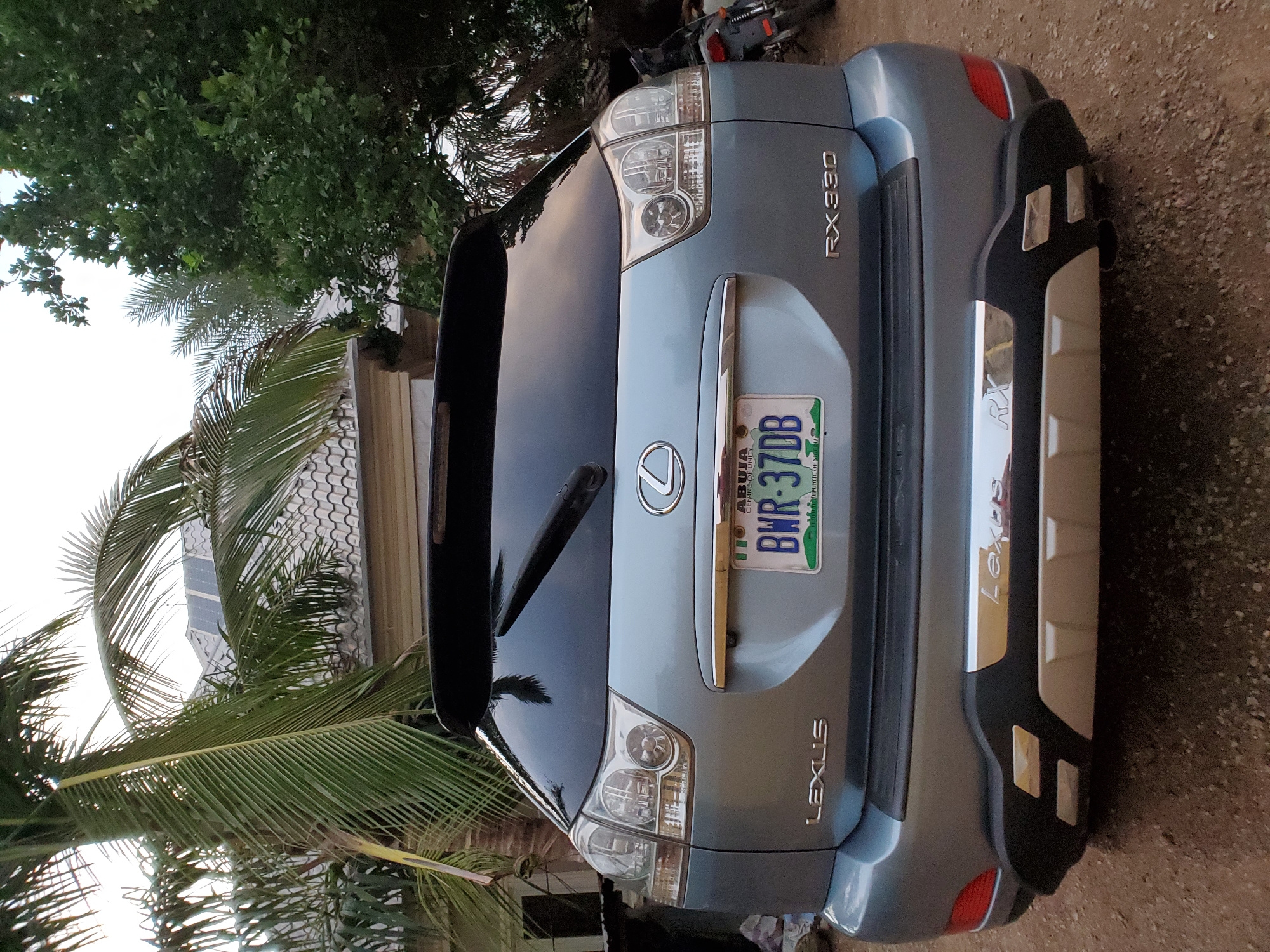 The image shows the rear of a silver Lexus RX330 SUV, license plate BWR-37DB, registered in Abuja, Nigeria. The car is parked outdoors on a dirt surface, with greenery and a building visible in the background. A motorcycle is partially visible above the car. The location is indicated by the license plate, but Mararaba is not mentioned, so the location is near Mararaba in Nigeria.