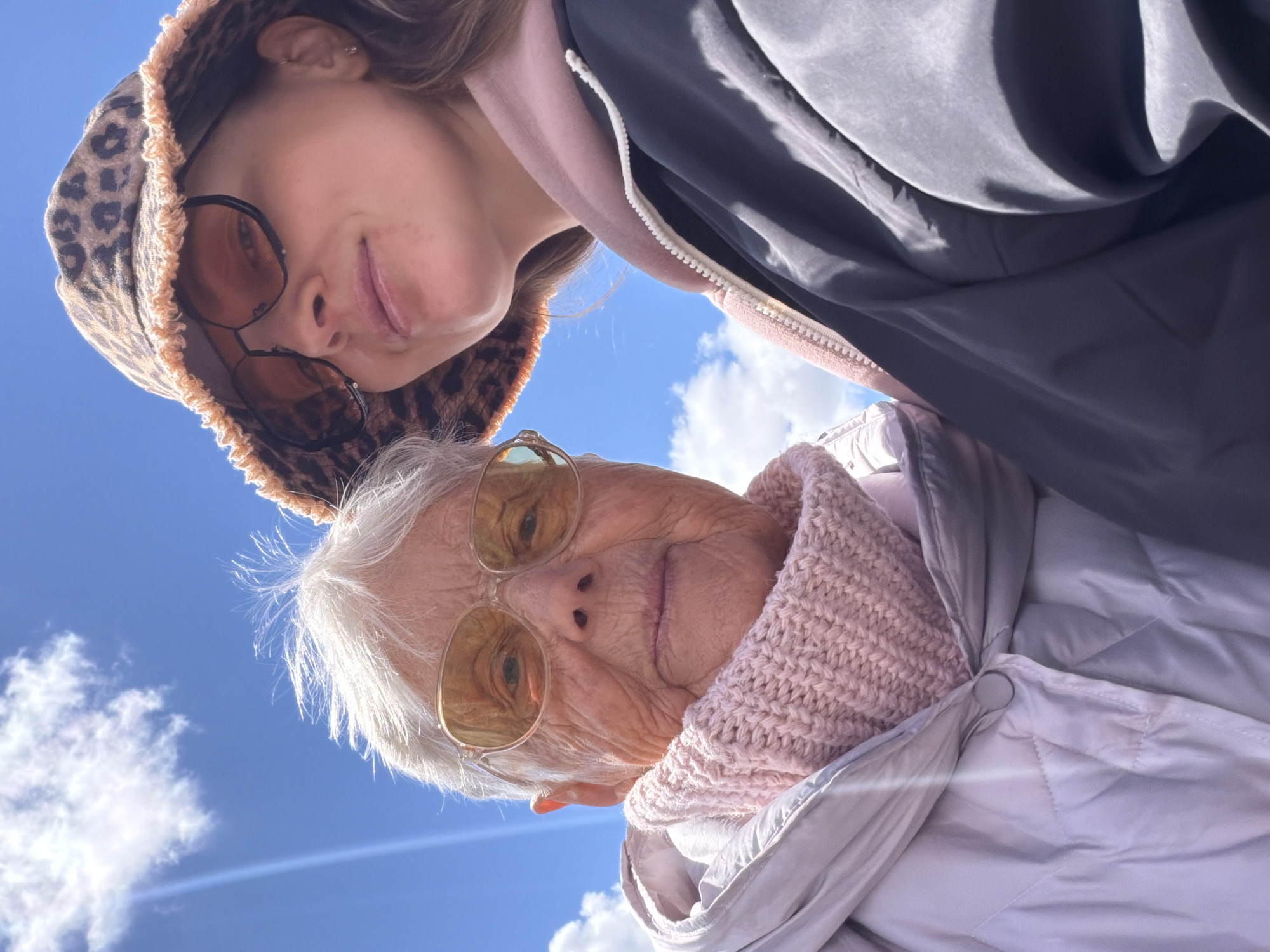 The image shows two people, likely related, standing close together outdoors. The younger person wears sunglasses and a leopard print hood, with a dark jacket partially visible. The older person wears sunglasses and a light pink knitted scarf with a light-colored jacket. Both individuals face the camera from a low angle. The background consists of a blue sky with scattered white clouds. There is no indication of the city of Valozhyn, Belarus.