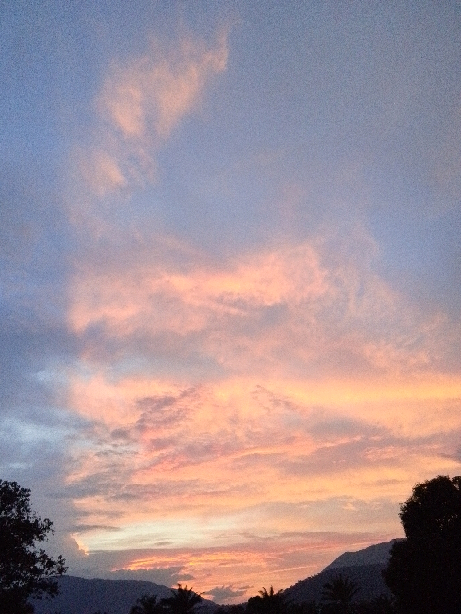 A panoramic shot captures the Serti, Nigeria sky at dusk, painted with a gradient of colors from soft blues at the top to vibrant pinks and oranges near the horizon. Wispy clouds, tinged with the warm hues of sunset, stretch across the expanse. Silhouetted against this dramatic sky are rolling hills and the dark shapes of trees and foliage, indicating a natural, possibly rural, environment on the outskirts of the city. The scene is devoid of any human presence or discernible activity, presenting a tranquil and serene landscape.