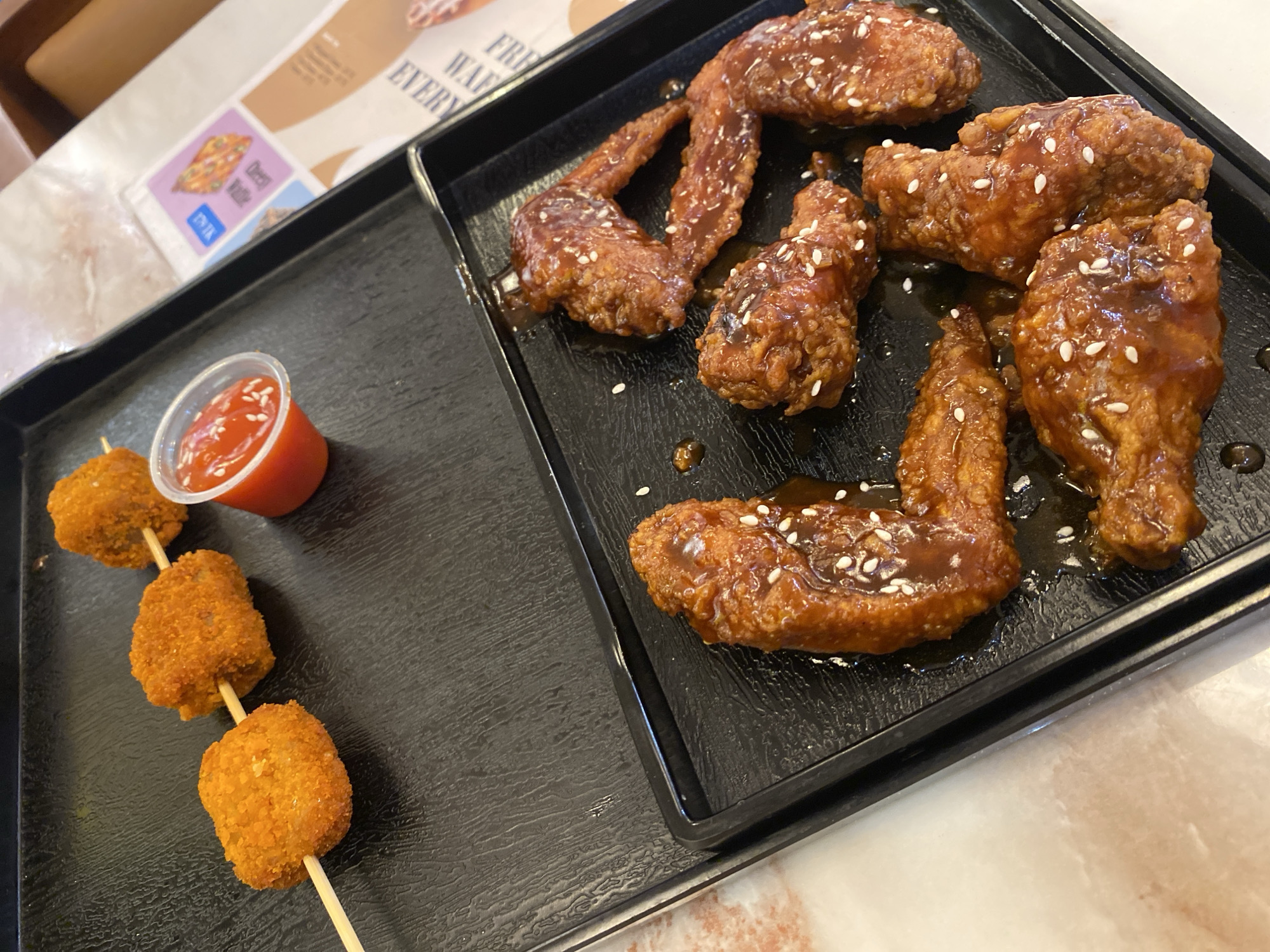 The image shows a black tray containing two dishes. On the left side is a skewer with three breaded, fried balls and a small plastic container of red sauce. On the right side are several pieces of fried chicken wings, covered in a dark sauce and sprinkled with white sesame seeds. The food is sitting on a table, presumably inside a restaurant. The location is Dhaka, Bangladesh.