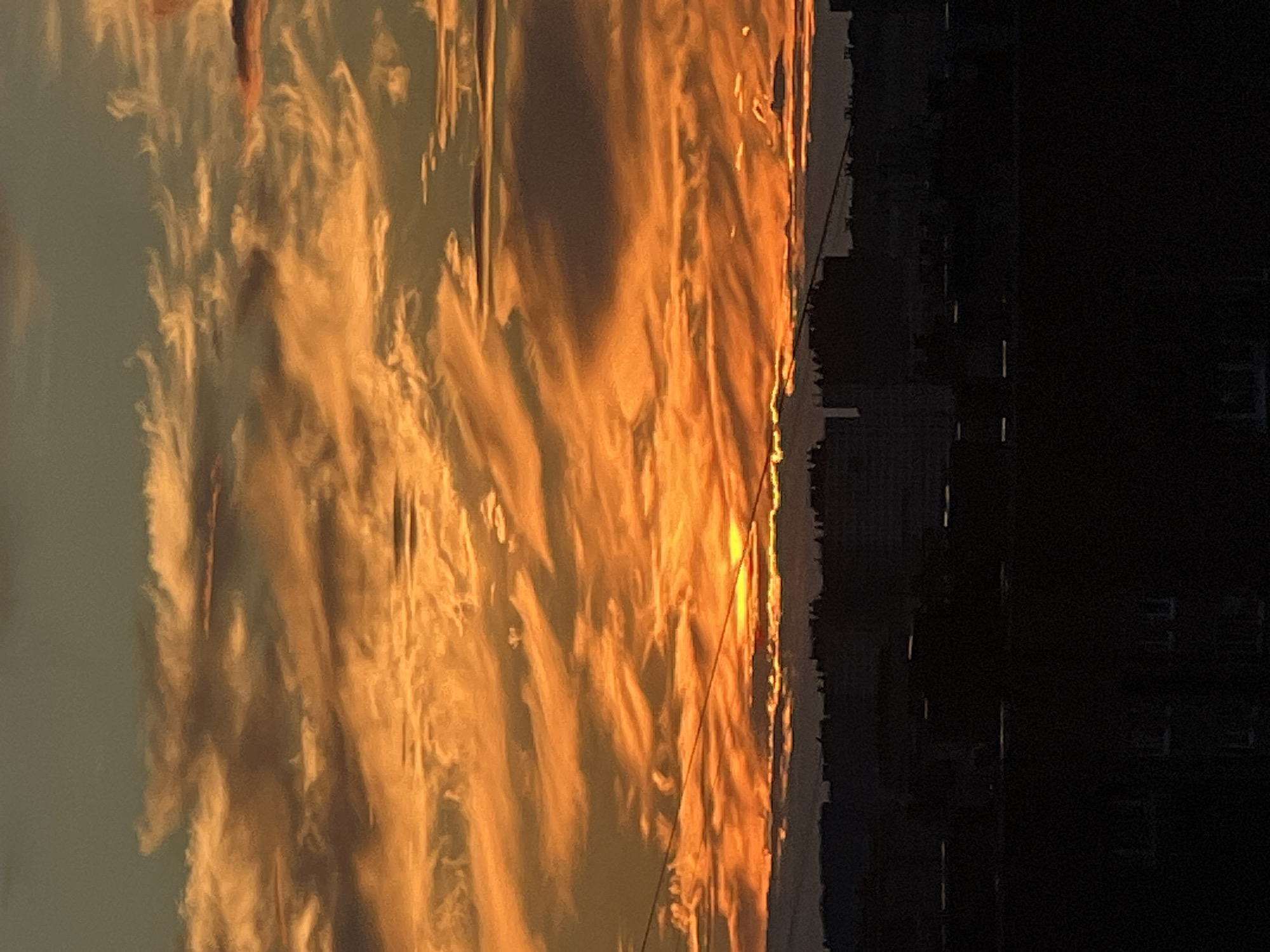 This media file depicts a dramatic sunset over a cityscape in Sinyavino, Russia. The sky is ablaze with vibrant orange and yellow hues, painted across wispy clouds that stretch across the frame. These fiery colors are reflected on a surface that appears metallic or glassy, creating a distorted and abstract interpretation of the sky. To the right of this reflective surface, the dark silhouette of buildings rises against the illuminated sky, hinting at an urban or suburban setting. The sun itself is visible as a bright, concentrated point of light on the horizon, just above the building line. The lighting suggests it is either sunrise or sunset, with the long shadows and warm tones being characteristic of these times of day. The overall mood is one of awe and perhaps a touch of melancholy, evoked by the intense natural beauty juxtaposed with the man-made structures. There is no visible text in the image.