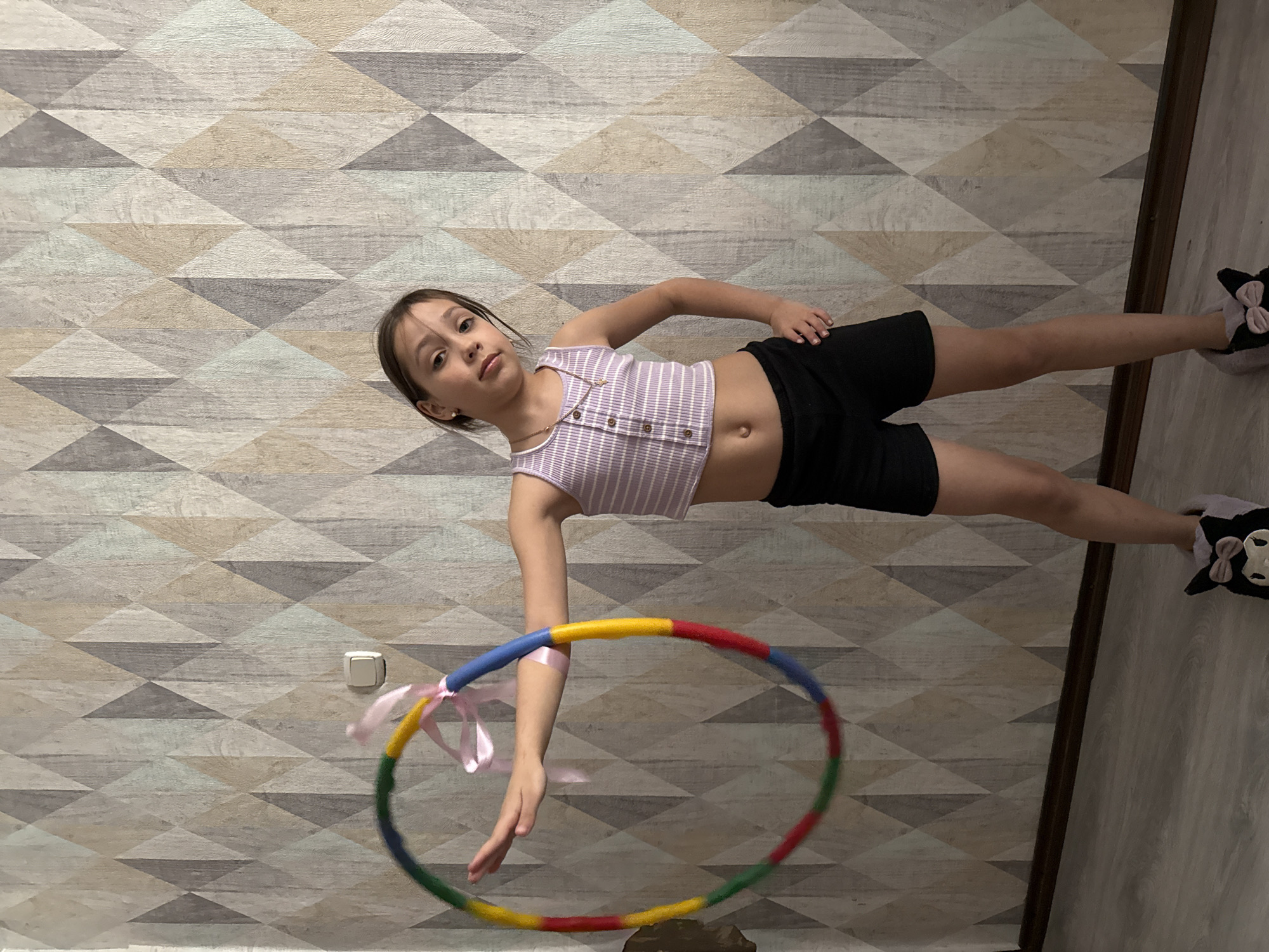 A young girl with brown hair and light skin poses in a room with geometric-patterned wallpaper. She wears a striped lavender tank top and black shorts, and has on black and white slippers. She stands with her left arm extended downwards, holding a colorful hula hoop with pink ribbons attached. The floor is wood-like on the right side of the frame. The image was most likely taken in an indoor setting, but the location in Votkinsk, Russia, cannot be determined from the image.