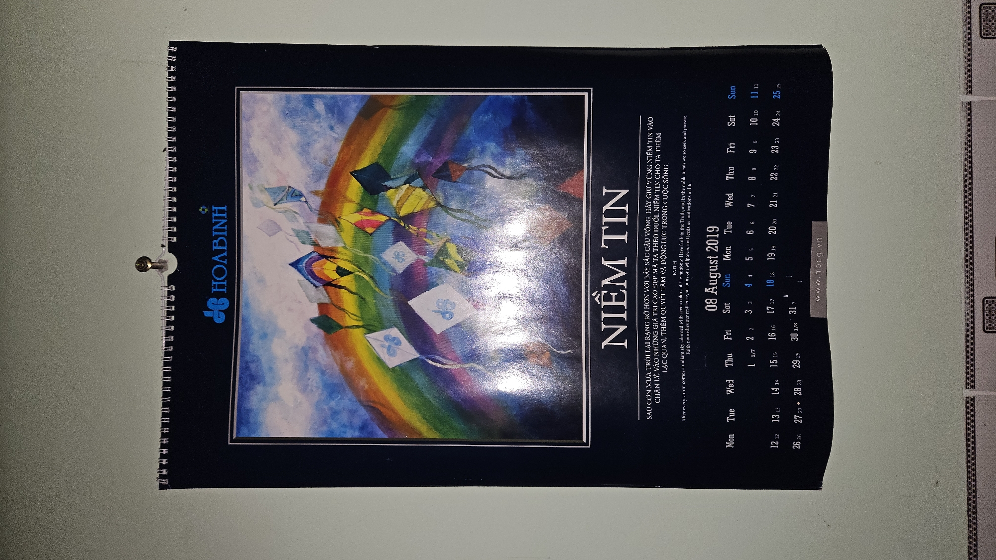 This media file displays a calendar page for August 2019, likely hanging on a wall in Cam Đức, Vietnam. The page is bound with a metal spiral, and a golden screw or nail for hanging is visible at the top left.
The upper portion of the calendar features a vibrant artistic illustration. The scene depicts a bright blue and purple sky adorned with a prominent, colorful rainbow. Numerous stylized kites, in various shapes and colors (yellow, orange, blue, green, white), are shown flying against this backdrop. Some kites feature abstract designs or symbols.
Below the artwork, the month and year "08 August 2019" are clearly visible. The calendar grid shows the days of the week (Mon to Sun) and dates from 1 to 31. Many of the dates are duplicated, possibly indicating days with specific events or holidays.
Prominently displayed in large, bold letters is the Vietnamese title "NIỀM TIN," which translates to "FAITH." Below this title, there is a motivational message presented in both Vietnamese and English. The English text reads: "After every storm comes a radiant sky adorned with seven colors of the rainbow. Have faith in the Truth, and in the noble ideals we so seek and pursue. Faith nourishes our resilience, sustains our willpower, and feeds us motivations in life."
On the left side, rotated vertically, is the company name "HÒA BÌNH" with its stylized "H" logo. At the bottom right, a website address "www.hbcg.vn" is visible. The overall aesthetic of the calendar, with its colorful artwork and uplifting message, conveys a sense of hope and inspiration.