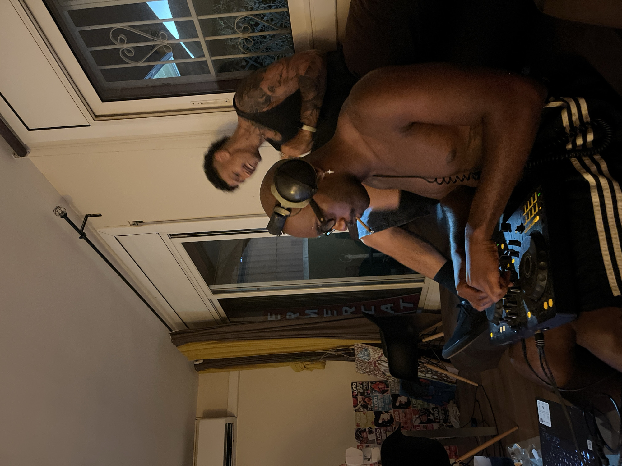 The image shows two shirtless men in a room. One man, wearing headphones and glasses, is operating a DJ mixing console. He is wearing shorts with stripes down the side. The other man, with tattoos on his arm, is standing behind him and smiling. A window with a metal grate is in the background, along with a sign that reads "MER CAT". The room appears to be in a residence, with a yellow curtain, air conditioning unit, and a wall decorated with posters. Based on the sign "MER CAT" visible in the window, the location is most likely in Barcelona, Spain. "Mercat" means market in Catalan.