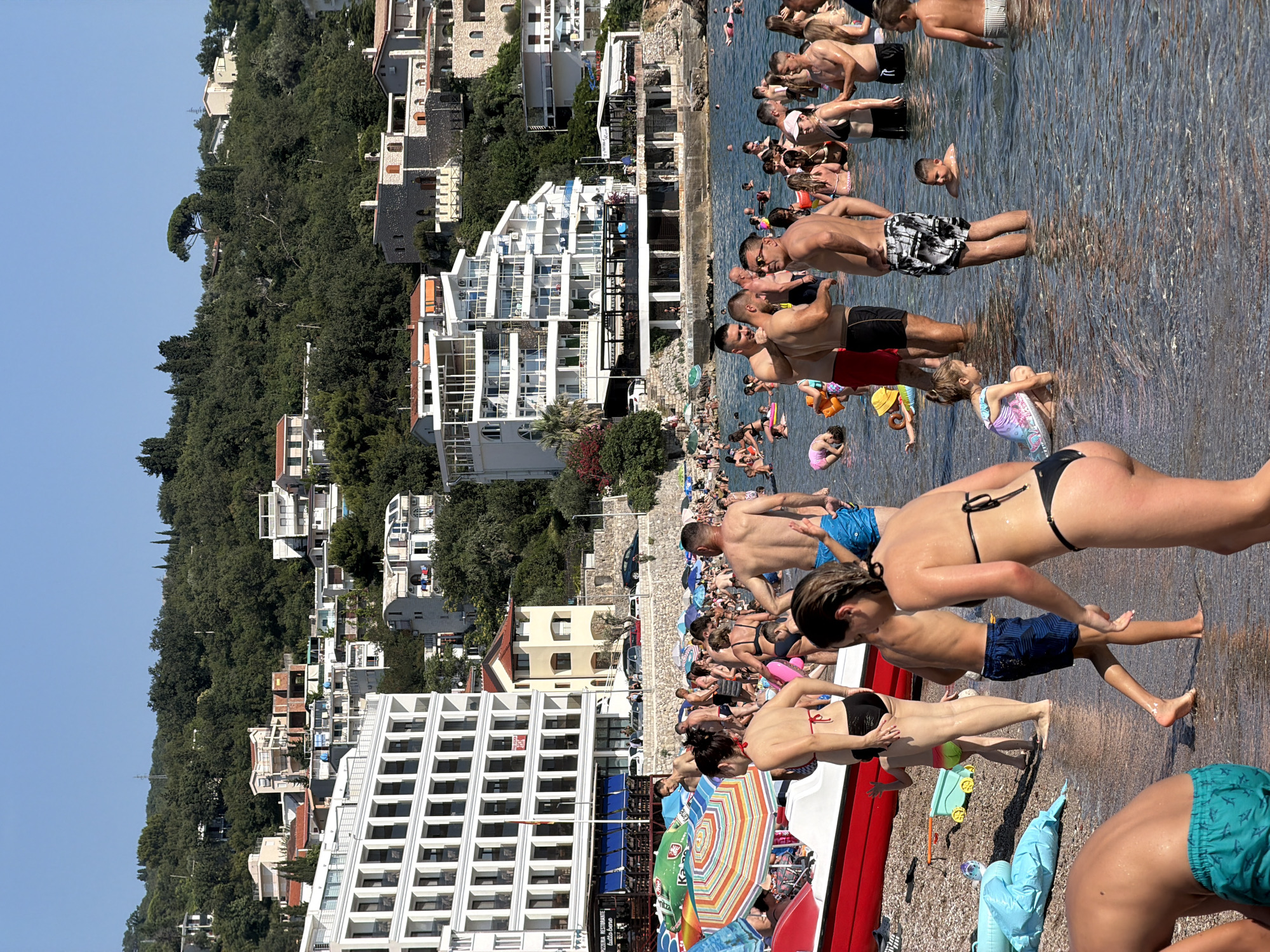 The image shows a crowded beach scene in Sutomore, Montenegro. Numerous people, including adults and children, are in the water and on the shore. The water is clear and shallow, with many individuals standing or wading. Beach umbrellas in various colors are scattered across the sand, along with beach toys and inflatable items. In the background, several multi-story white buildings with balconies line the coast, and further inland, houses are built on a lush, green hillside under a clear blue sky. The scene suggests a lively and popular tourist destination during daylight hours.