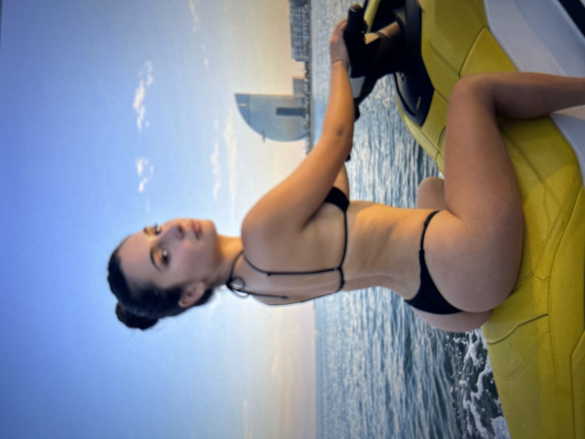 A young woman with her hair pulled back in a bun, wearing a black bikini, is riding a yellow jet ski on the sea. She is looking back at the camera. In the background, a distinctive sail-shaped building, the Hotel Barcelona, is visible along the coastline, suggesting the location is Barcelona, Spain, not Burgas, Bulgaria. The sky is a clear blue with a few wispy clouds, and the sun is low, casting a warm light on the scene. The water is calm with some small waves.