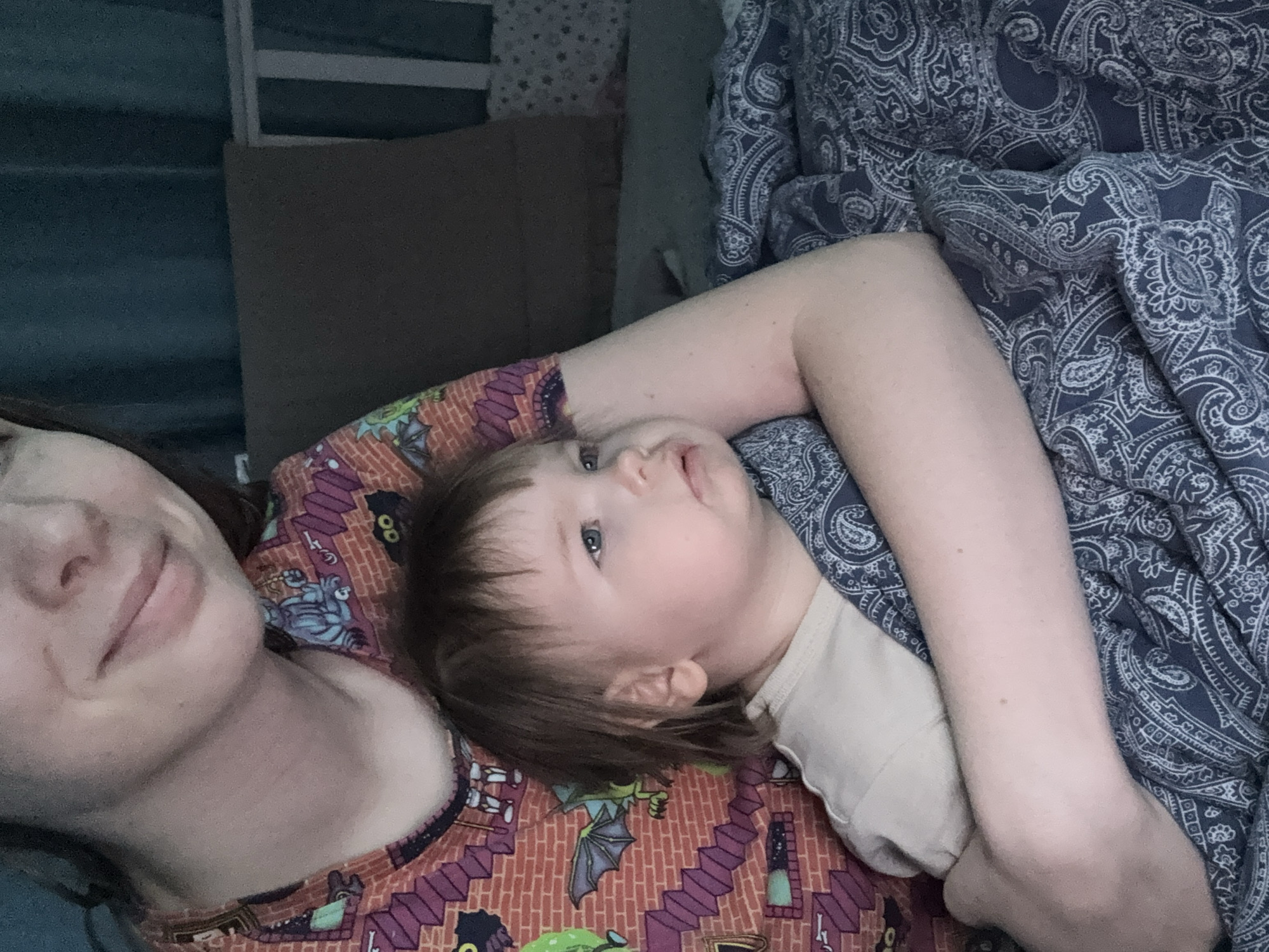 A mother and child are lying together on a bed. The mother is on her left side with the child nestled on her chest. The mother is wearing an orange shirt with Halloween-themed designs. The child is wearing a light-colored shirt. A blue paisley patterned blanket is draped over the child's body. A brown pillow is partially visible behind the mother. The location appears to be indoors. There is no indication of Moscow, Russia.