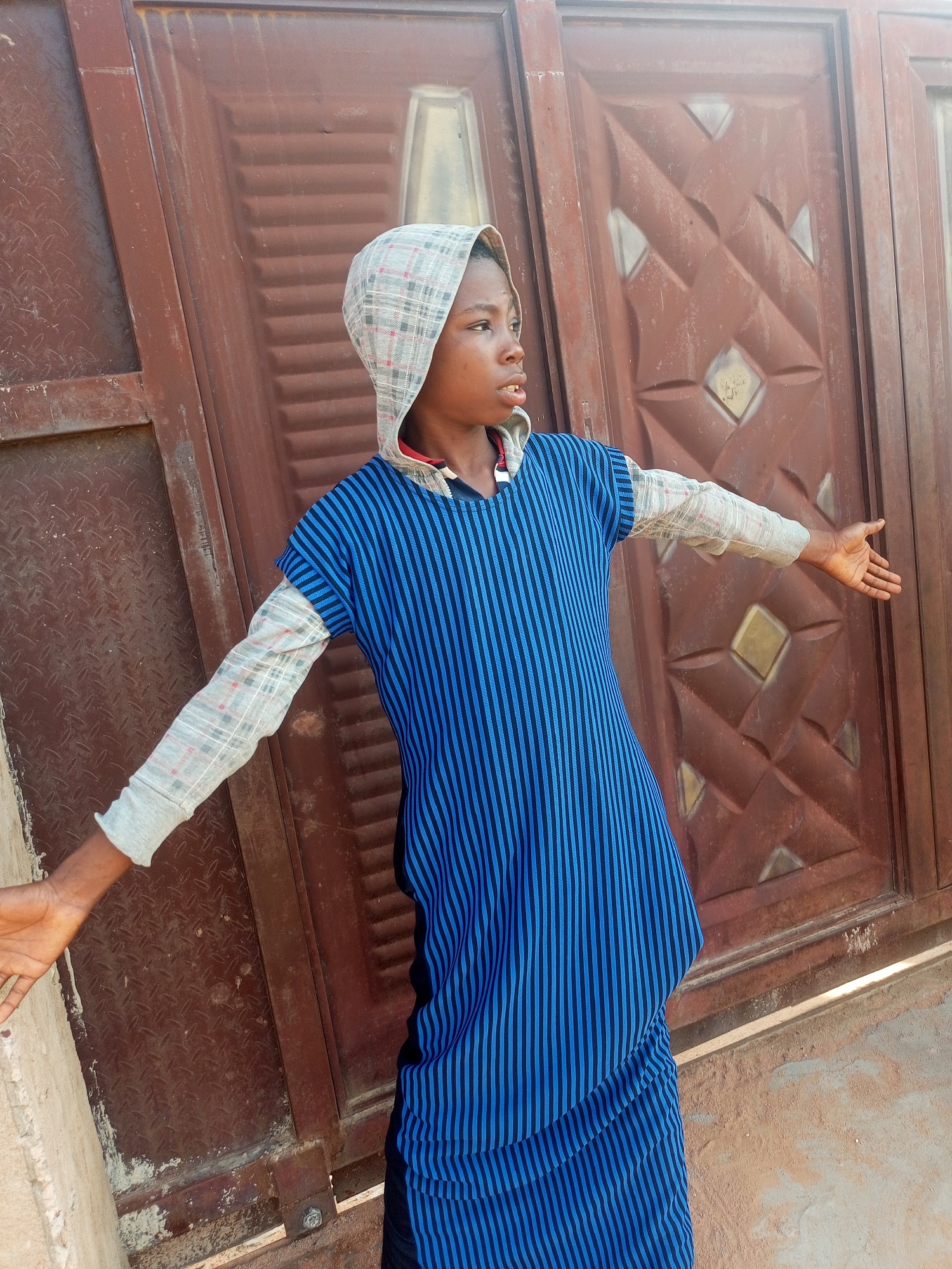 A young boy stands with his arms outstretched in front of a dark brown, weathered metal gate. The gate has a diamond-patterned design. The boy is wearing a hooded garment with a checkered pattern under a blue and black striped tunic. His left arm is extended to the left, and his right arm is extended to the right, with his palms open. The background is dominated by the gate, with a portion of a textured, light-colored wall visible on the left side of the frame. The scene appears to be outdoors, possibly in the city of Tempure, Nigeria.