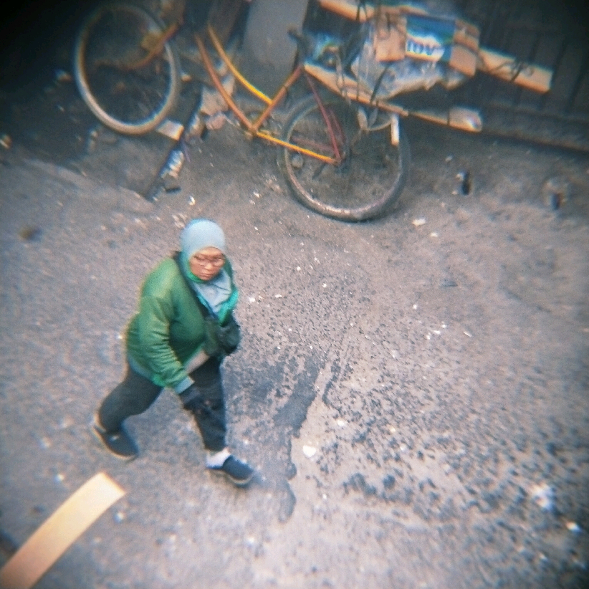 The image shows a person, likely a woman, from a high-angle perspective, wearing a light blue hijab and a green jacket. She is walking on a paved surface that appears to be a street or sidewalk. Her clothing and the general setting suggest a temperate or warm climate. To the left of the person, a bicycle is partially visible, with its wheels and frame suggesting it is an older model, possibly used for carrying goods given its sturdy build. There is some indistinct text visible on the bicycle's cargo area, with "AGI" being the most legible part. The overall scene is somewhat blurry, which could be due to motion or the quality of the original photograph, but it conveys a sense of everyday life in an urban environment. The lighting suggests it could be daytime, though the exact time is not discernible. The ground texture appears rough, typical of city streets in some regions. The person's posture and focus suggest they are engaged in an activity such as commuting or running errands. The presence of the bicycle implies it might be a mode of transportation or a tool for work. The overall impression is one of candid street photography capturing a moment in time.