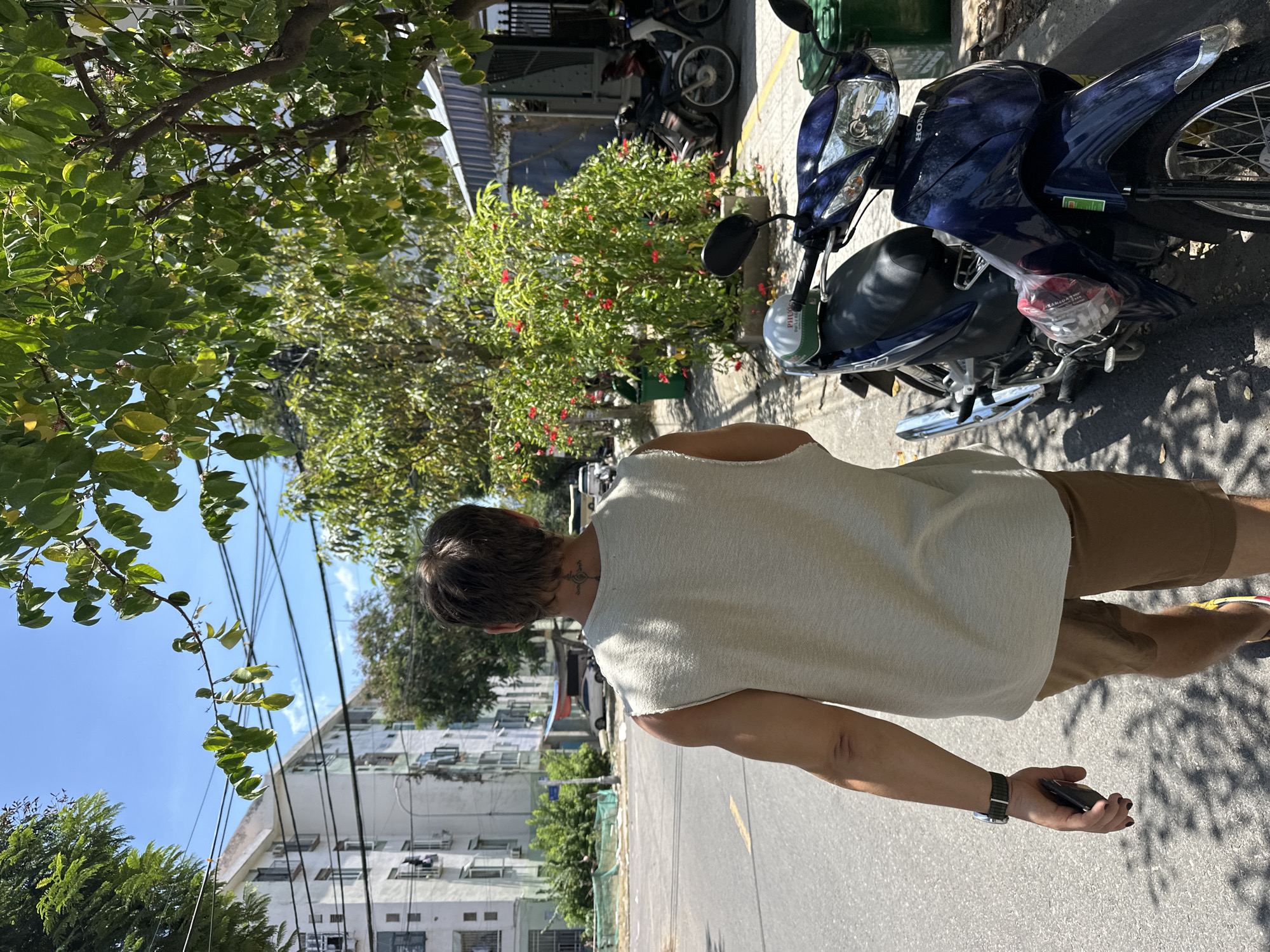 A person with a tattoo on their neck is walking away from the camera on a city street in Da Nang, Vietnam. They are wearing a sleeveless off-white top and brown shorts. A dark blue motorcycle is parked on the right side of the street next to a green trash can. In the background, there are apartment buildings, trees with red flowers, and other parked motorcycles. The sky is blue with some clouds. The person is holding a black mobile phone in their left hand.