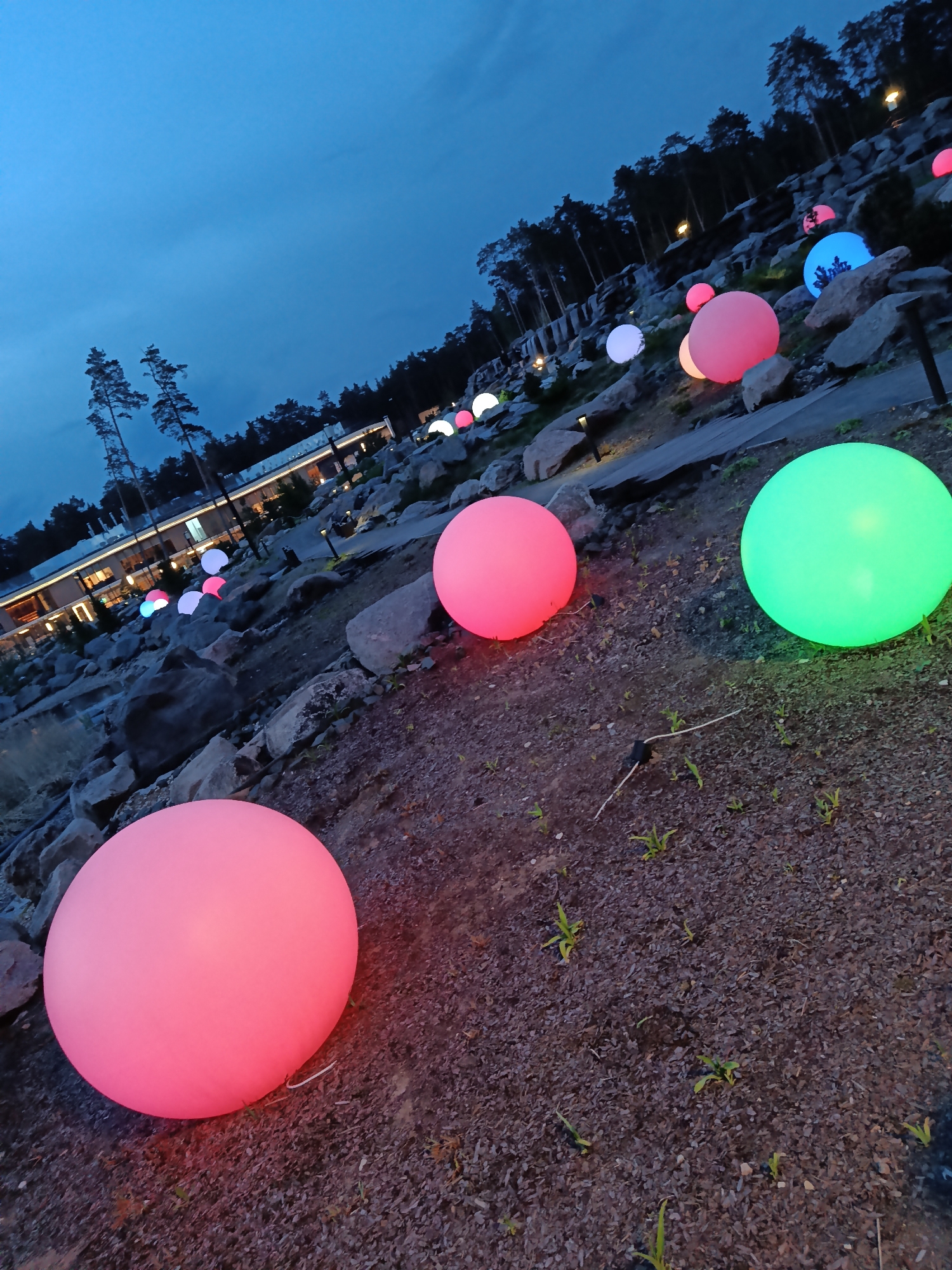 The scene is a landscaped area in Shulgino, Russia, at dusk. Large, illuminated spheres in pink, green, white, and blue are scattered across the ground and among rocks. A building with lighted windows is visible in the background, surrounded by trees. The ground is covered with dark gravel or soil, with sparse patches of vegetation. The lighting creates a whimsical, festive atmosphere.