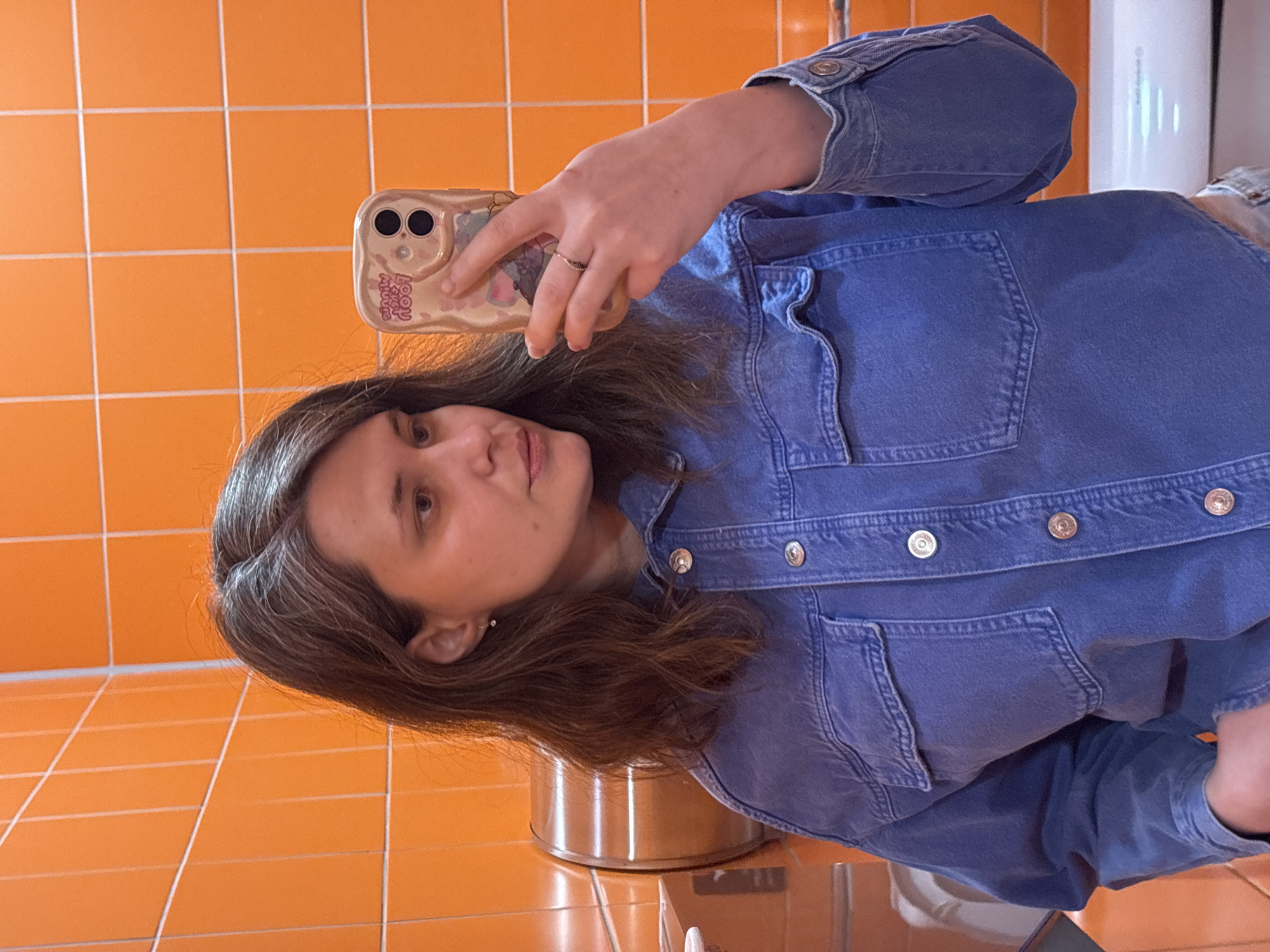 An adult female is depicted taking a mirror selfie in a tiled room, identified as being in Izhevsk, Russia. She has medium-length brown hair with visible graying strands, styled with a side part. She wears a blue denim shirt or jacket featuring light-colored buttons and two chest pockets. A small, clear stud earring is present in her visible right earlobe. Her right hand holds a light-colored smartphone with a two-lens camera visible, enclosed in a reflective, patterned case that displays inverted text, including "MINNIE" and "POOH". A ring is present on her right ring finger. She gazes slightly to her right. The background consists of bright orange square wall tiles with white grout lines. To the left of the subject, a cylindrical, reddish-brown metallic fixture, possibly a hand dryer or dispenser, is mounted on the wall. A white porcelain fixture, likely a toilet or urinal, is partially visible in the lower right corner of the frame.