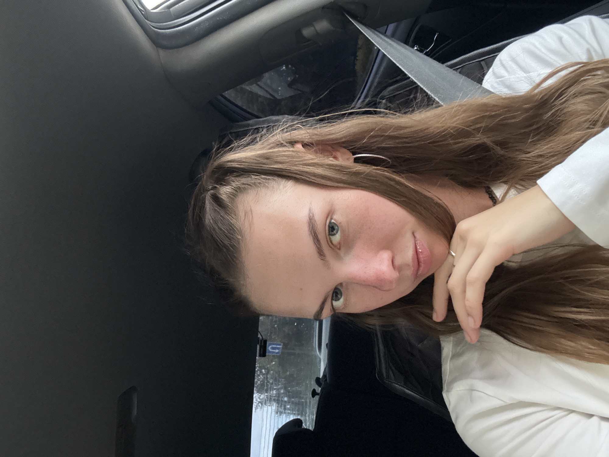 A young woman with light brown hair is shown from the chest up, sitting in the passenger seat of a car. She is looking directly at the camera with a neutral expression. She is wearing a white shirt and a silver hoop earring in her left ear. Her right hand is resting on her chin, and she is wearing a silver ring on her right ring finger. The background shows the interior of the car, including the headrest of the seat in front of her and a seatbelt. The window behind her shows a blurred view of trees and a street sign with a "U-turn" symbol. The lighting suggests it is daytime. The location is the city of Perm, Russia.