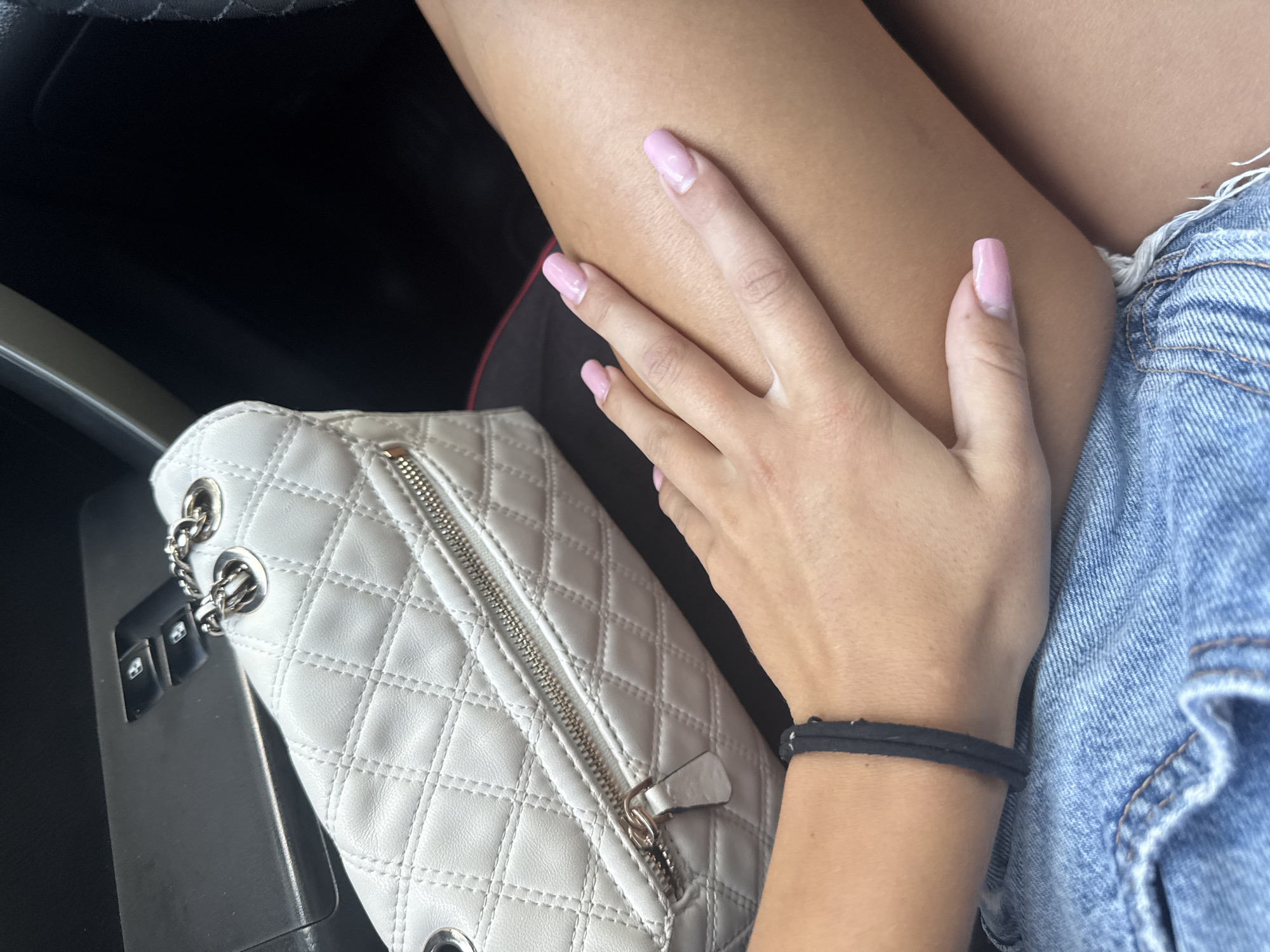 In Basarabeasca, Moldova, a close-up shot captures a person's left hand, adorned with light pink manicured nails, resting on their thigh. The person is wearing denim shorts. Beside the thigh, a light-colored, quilted handbag with silver chain accents is visible. In the background, a car's interior is partially seen, including what appears to be a central console with buttons. The lighting suggests it is daytime, possibly indoors or in a shaded outdoor area.