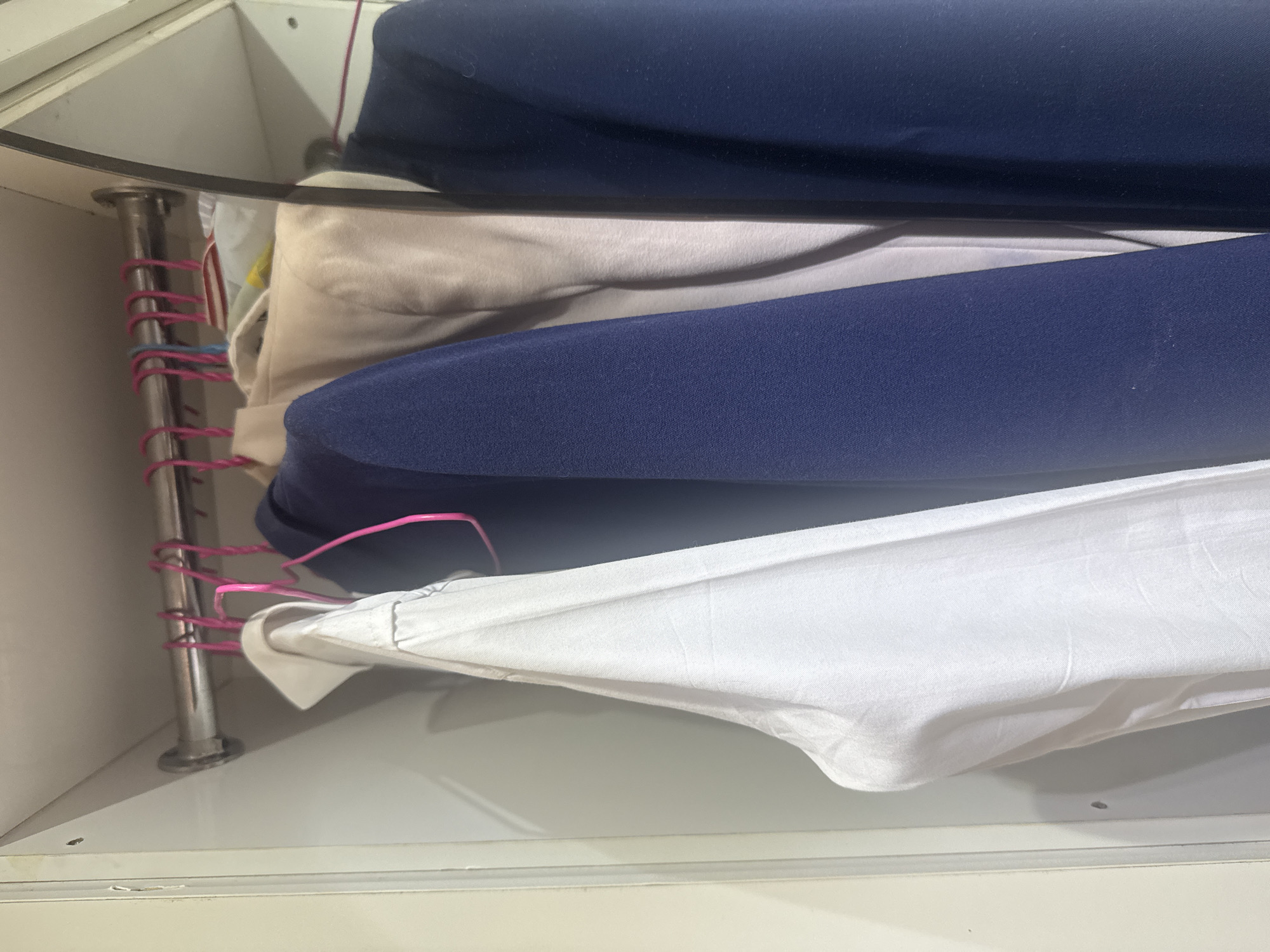 The image shows a close-up view of folded clothing inside a closet or storage unit. The clothing consists of several layers of fabric in different colors, including dark blue, beige, and white. The items appear to be folded neatly and stacked on top of each other. A metal rod, possibly a shelf support or part of a hanging mechanism, is visible on the left side of the image. Pink-colored wires or cords are wrapped around this rod, adding a splash of color to the otherwise neutral palette of the clothing and storage unit. The lighting suggests an indoor setting, with no strong shadows indicating a specific time of day. The background is out of focus, but it appears to be a plain, light-colored surface, possibly the interior of the closet. There is no visible text in the image. The overall impression is one of organized storage. The context provided is Abuja, Nigeria, but this information cannot be visually confirmed from the image itself.
