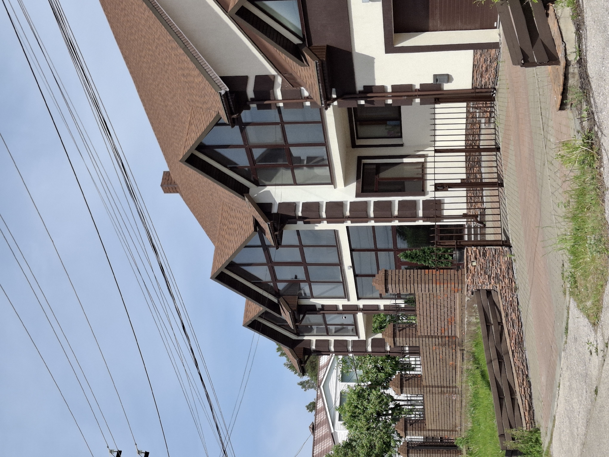 The image shows a multi-story building in the Прикамский region of Russia. The building features a brown roof, white exterior walls, and large windows with brown frames. A metal fence runs along the perimeter of the property. The sky is visible in the background, along with power lines. A paved walkway and some greenery are also visible.