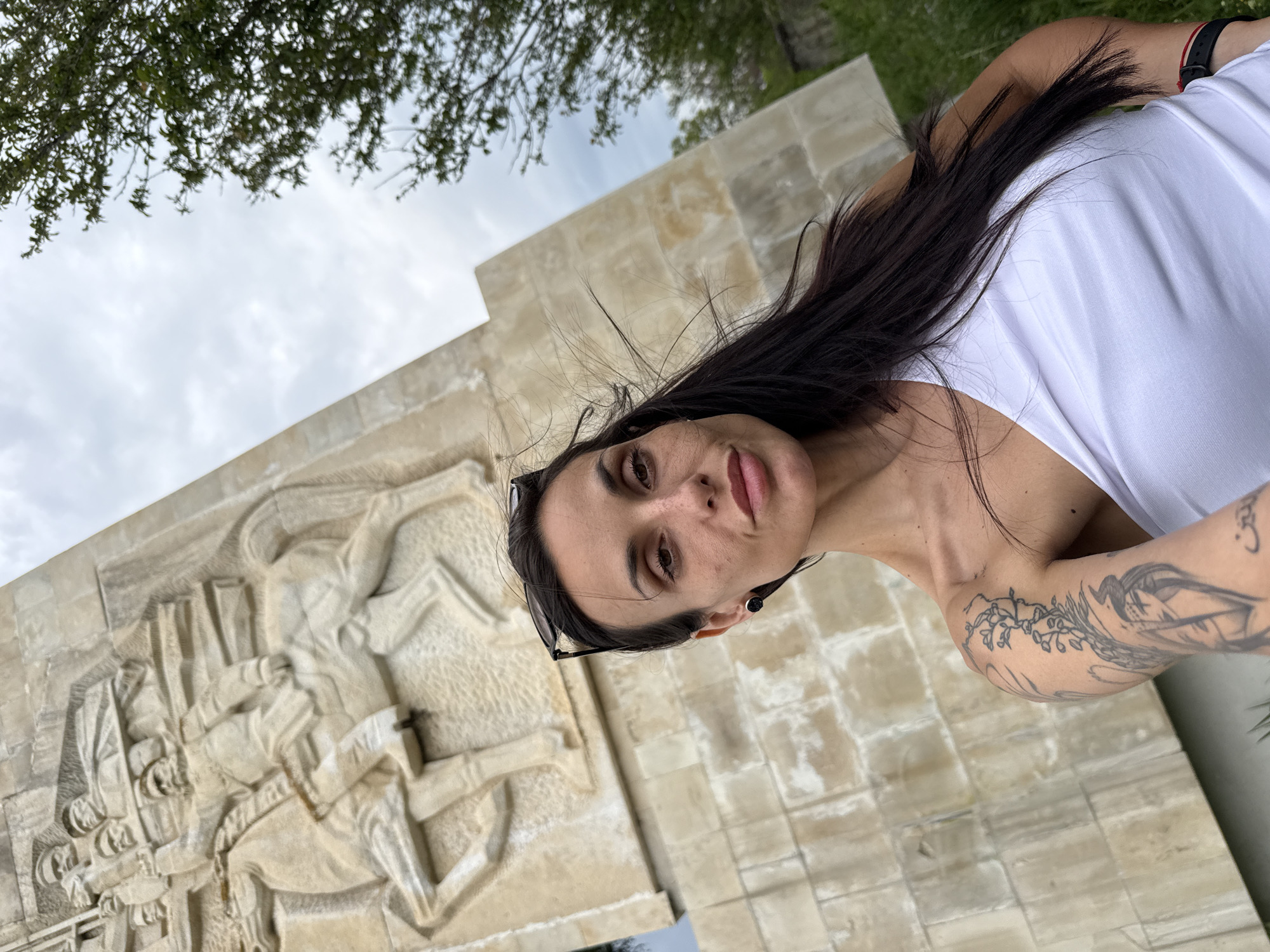 This close-up, tilted image captures a woman taking a selfie or being photographed outdoors, with a substantial historical monument in the background. The woman, positioned on the right, has long, dark hair, dark eyes, and full lips. She is wearing a white sleeveless top and has sunglasses pushed up onto her head. Distinctive tattoos, including a floral or vine-like pattern and other stylized designs, are visible on her left arm and shoulder. A black wristwatch is on her right wrist. Her expression is neutral, with her lips slightly parted.
The background features a large, light-colored stone monument, possibly made of limestone or marble. It is adorned with intricate relief carvings depicting multiple figures in historical attire, suggesting a scene of battle or a significant historical event. The monumental structure fills a large portion of the left and center background.
The setting is an outdoor area with natural elements. Dark green foliage from trees is visible at the top left of the frame, and patches of green grass can be seen. The sky is overcast with soft grey clouds, indicating a cloudy day without harsh sunlight. Given the location context of Pleven, Bulgaria, the monument is strongly suggestive of the Panorama "Pleven Epopee 1877" or a related memorial commemorating the Russo-Turkish War. There is no visible text in the image beyond the graphical elements of the tattoos.