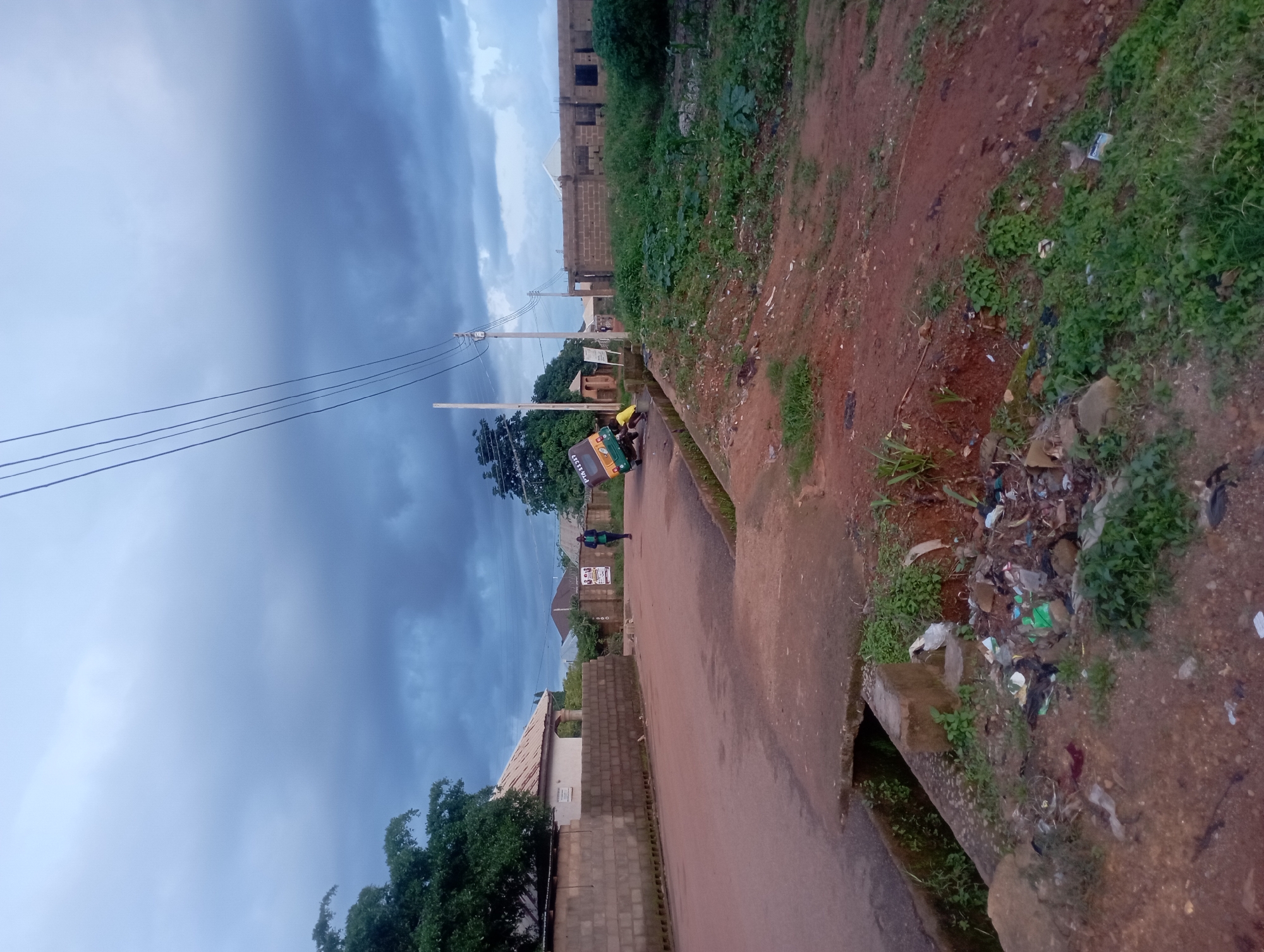 The image depicts a residential street scene in Rafin Bauna, Nigeria, under a cloudy sky. In the foreground, a pile of accumulated garbage lies on the reddish-brown dirt to the right of a concrete drainage channel. The channel runs along the edge of a paved road that curves to the left. A person in a dark jacket and light pants is walking away from the camera on the road. Further down the road, a yellow and green auto-rickshaw is overturned on its side, with two individuals, one wearing a yellow shirt, attending to it. A utility pole with multiple wires stands on the left side of the road. Beyond the road, on the left, a single-story building with a corrugated metal roof is visible behind a brick wall. On the right side of the road, construction of a two-story brick building is underway. Lush green vegetation is present throughout the scene, with trees on the left and denser foliage on the right, particularly around the construction site. The overall atmosphere is somewhat somber due to the overcast weather.