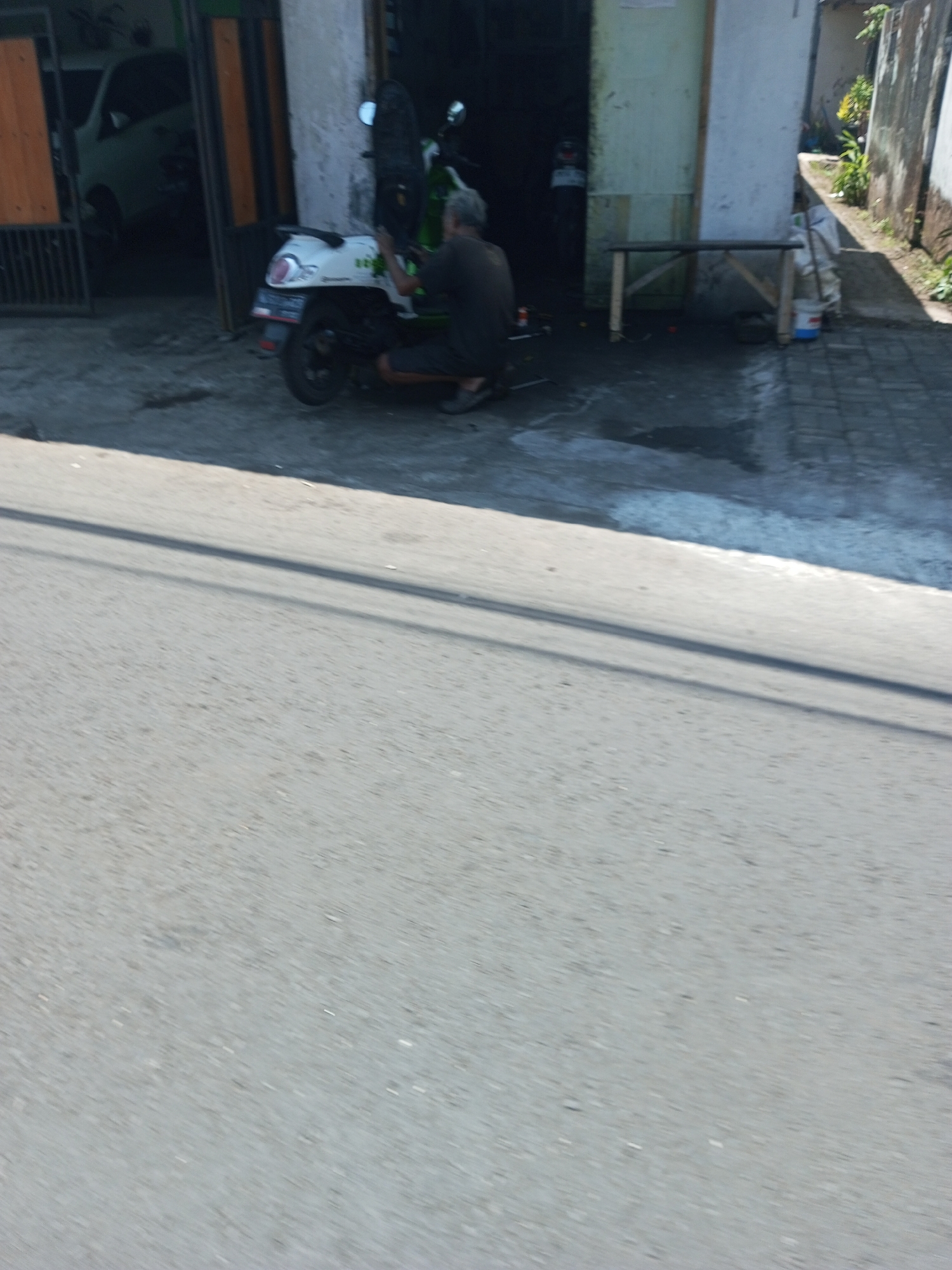 A person is kneeling beside a white scooter outside a building in Purwosari Klampok, Indonesia. The person, with gray hair, wears a dark shirt and shorts. The scooter is parked on a gray paved surface. Behind the scooter and the person is a building with an open doorway revealing a white car inside. A wooden bench is positioned to the right of the doorway. The ground outside the building is paved with gray bricks.