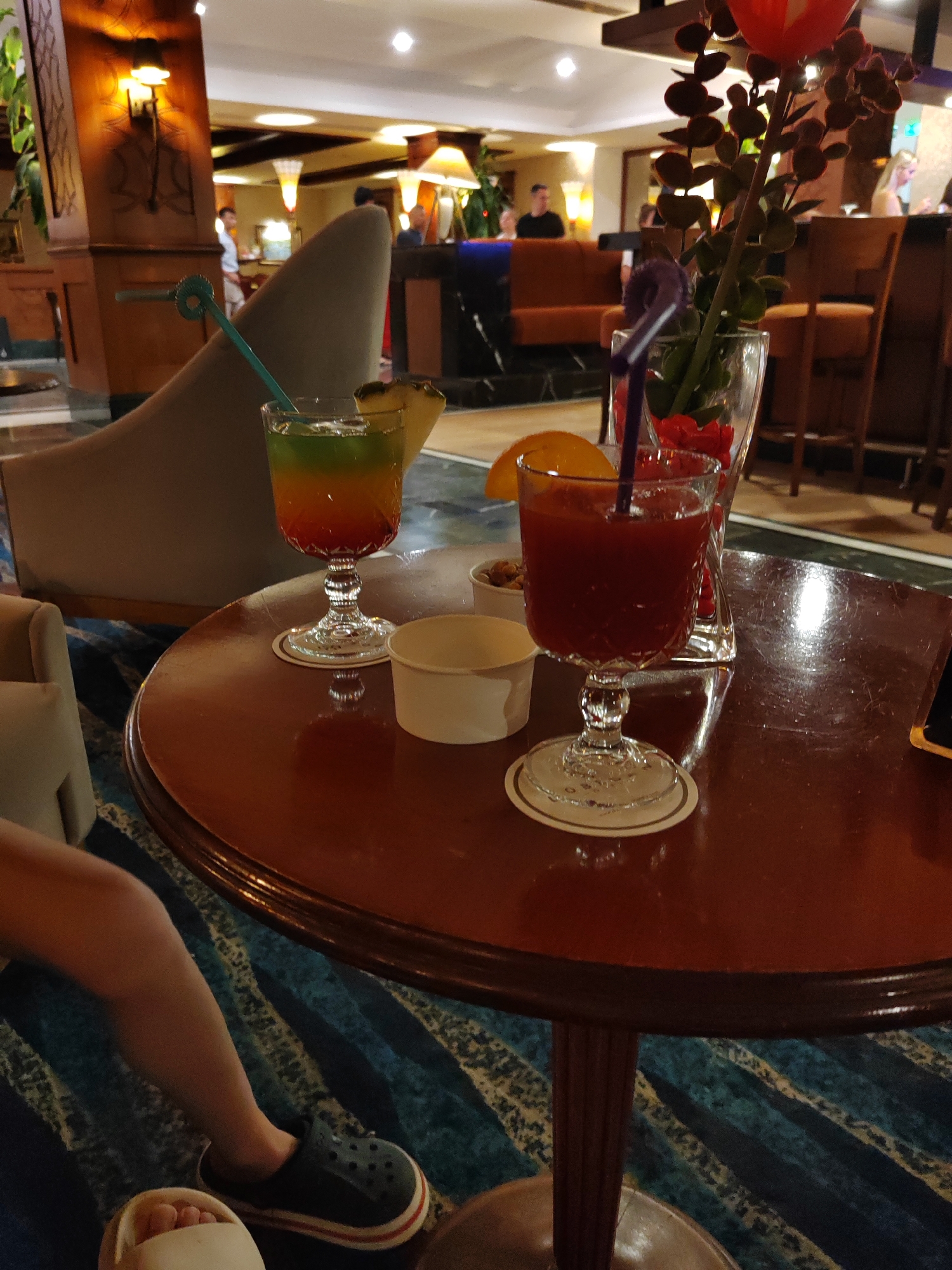 The image shows a close-up view of a round, dark wooden table in what appears to be a hotel lobby or lounge area. On the table, there are two cocktails. The cocktail on the left is a layered drink with red, orange, and green hues, served in a stemmed glass and garnished with a slice of pineapple. A blue-green straw is sticking out of this drink. To the right of this glass, there is a small white paper cup, and further to the right, another cocktail is served in a stemmed glass, this one appearing to be a deep red color, with a straw and a slice of orange. Behind the glasses, a clear vase holds a decorative artificial plant with red accents and a purple straw. In the background, blurry figures of people can be seen, some standing and others seated, in what looks like a bar or seating area with orange seating. The lighting suggests an indoor evening setting, with warm-toned lamps visible. The floor is covered in a patterned blue and white carpet. The presence of multiple seating areas, the bar setup, and the general ambiance point to a hotel or resort in Manavgat, Turkey, likely a common area where guests relax and socialize. A person's legs and feet are visible in the lower left corner of the image, wearing dark clogs and white slippers, suggesting they are seated at or near the table.