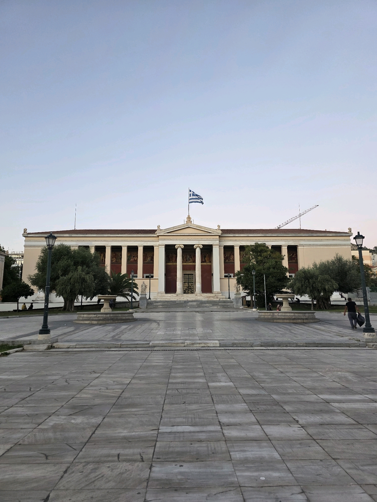 The image shows the exterior of the National and Kapodistrian University of Athens. The building is a neoclassical structure with a colonnade, a pediment, and a Greek flag flying at the top. In front of the building is a paved courtyard with trees, a fountain, and streetlights. A single person is walking away from the camera towards the right side of the frame, carrying a bag. The sky is clear and pale blue, suggesting it is either early morning or late evening. The overall scene is tranquil and depicts a well-maintained public space.