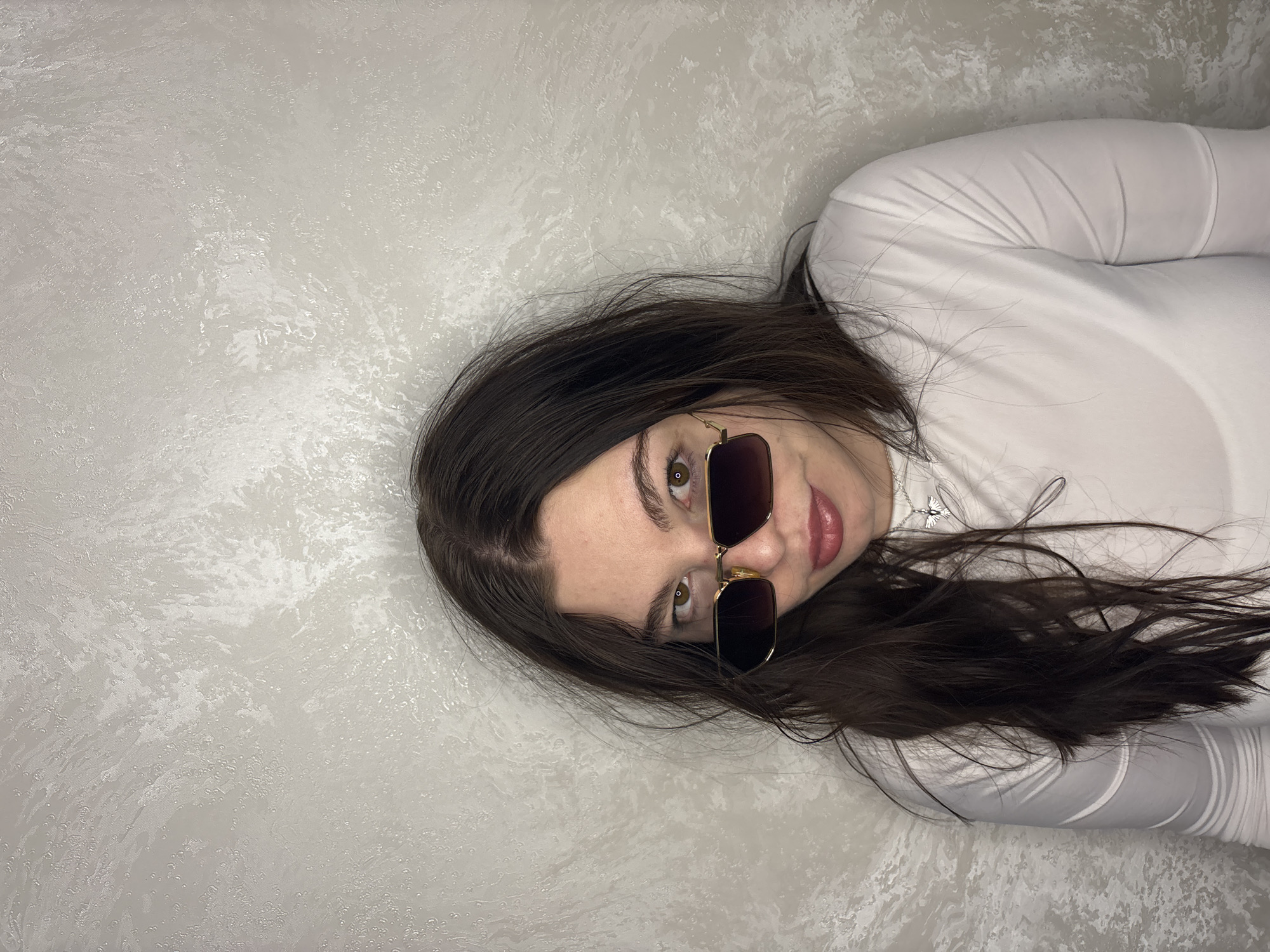 A woman with long dark brown hair and brown eyes, wearing gold-framed sunglasses, lies against a textured, light grey surface. She is wearing a white, long-sleeved shirt. The image may have been taken indoors against a wall or on a surface with a patterned texture. There is no clear evidence to indicate that the photograph was taken in Perm, Russia.