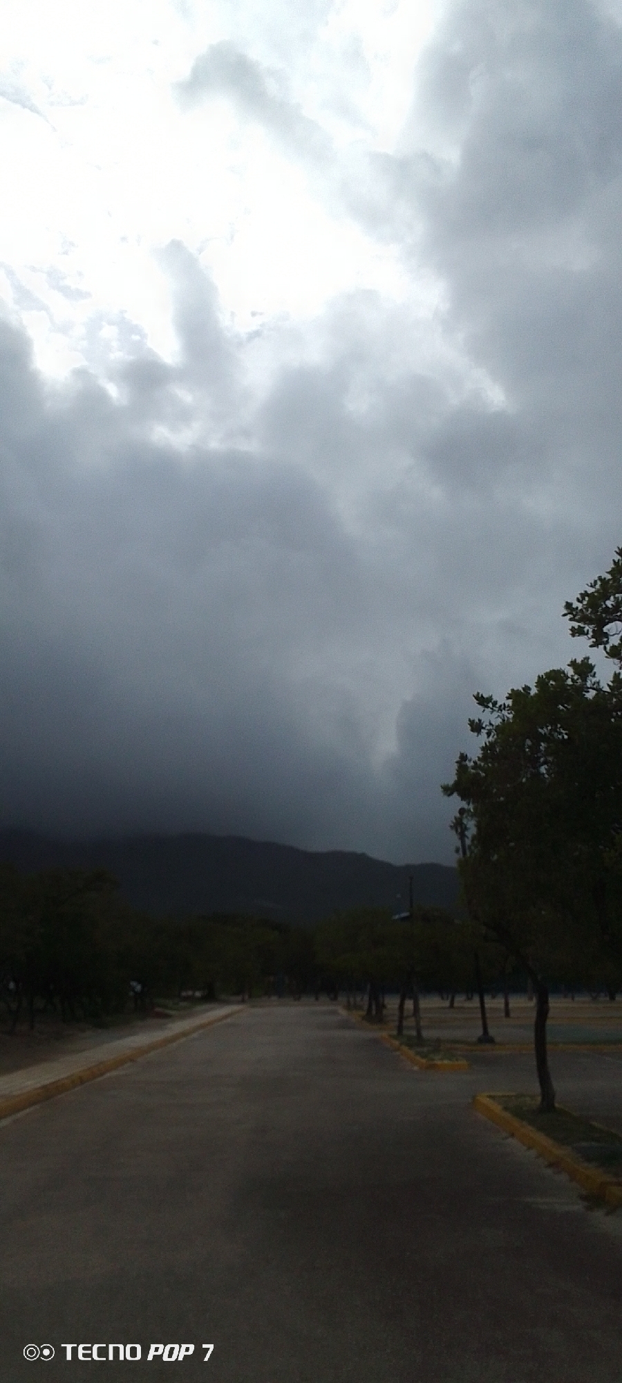 This media file depicts an outdoor scene, likely a parking lot or road adjacent to a park or recreational area, under a heavy, overcast sky. The foreground shows a paved surface, possibly a road or parking aisle, with a distinct yellow border on the left and right edges. Rows of trees, sparsely leafed, line the sides of the paved area, suggesting a dry season or a particular type of vegetation. In the background, a dark, mountainous silhouette is visible against the dramatically clouded sky, hinting at a large natural landscape. The overall mood is somber and perhaps slightly ominous due to the dark, dense clouds, indicating a potential for rain or a storm. The time of day appears to be late afternoon or early evening, given the dim lighting and the color of the sky. The location is identified as Pedro González, Venezuela. The bottom left corner of the image displays the visible text "© TECNO POP 7". There are no people or specific activities clearly depicted; the focus is on the landscape and atmospheric conditions.