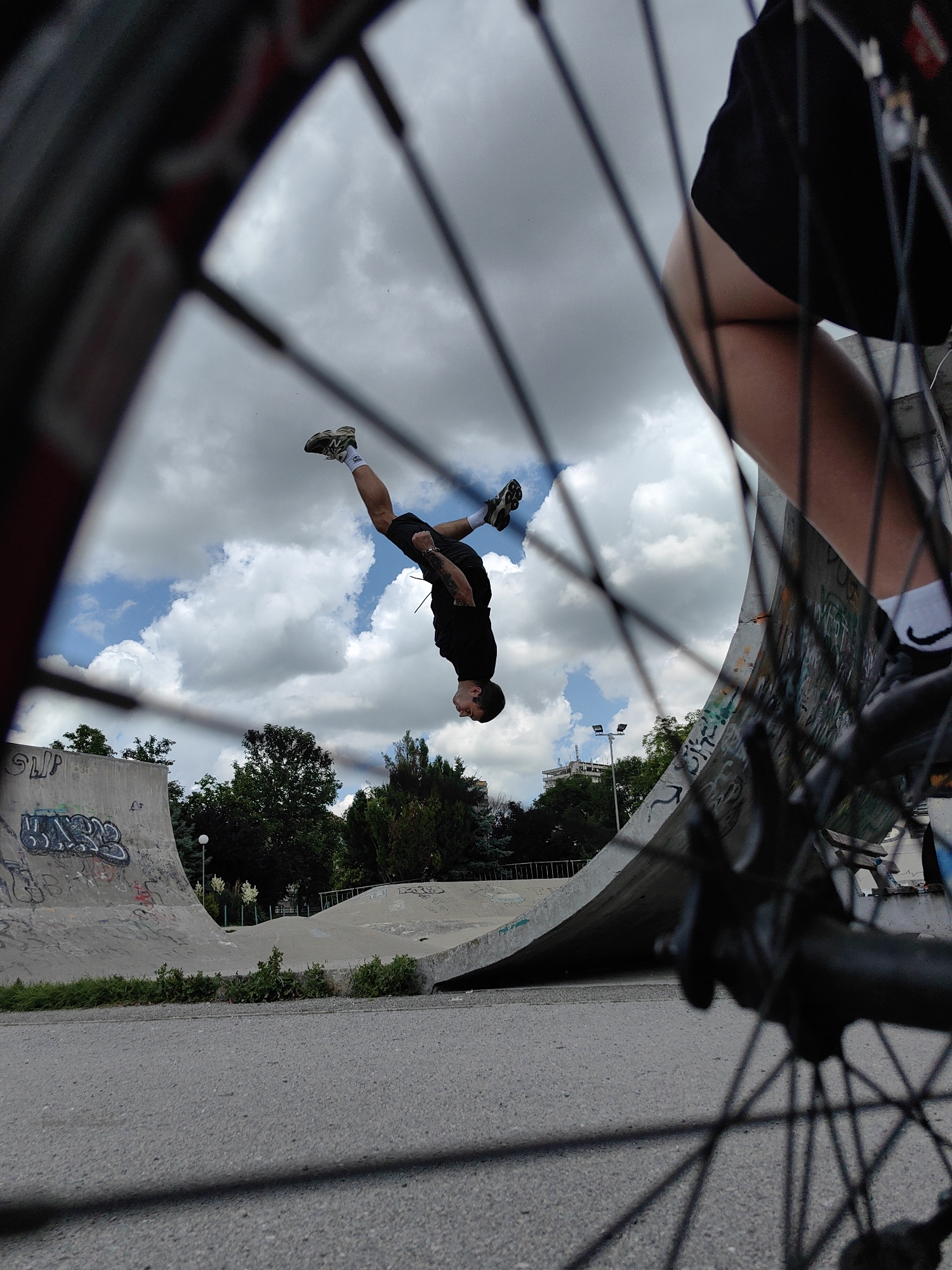 The image shows a young man performing a backflip in a skate park. He is captured mid-air, upside down, with his legs extended. He is wearing a black t-shirt, black shorts, and athletic shoes. The skate park appears to be concrete with various ramps and structures, some of which are covered in graffiti. In the foreground, the spokes of a bicycle wheel are visible, framing the scene. The sky is partly cloudy with blue patches. The location is Shumen, Bulgaria.