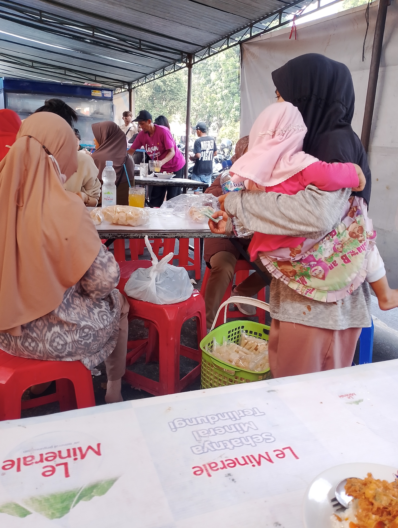 The image depicts an outdoor dining scene under a covered structure, possibly a food stall or restaurant, located in the city of Yogyakarta, Indonesia. The environment is casual, with patrons seated at tables.
In the foreground, a person wearing a light brown hijab and a patterned dress is seated on a red plastic stool. In front of them, a white plastic bag containing food is placed on another red plastic stool. A white plastic table is visible, laden with food items, including what appear to be bags of snacks and a bottle of water. A glass of orange beverage is also present.
In the mid-ground, a woman wearing a black hijab is holding a young child. The child is wearing a pink headscarf and a pink shirt, with a printed cloth draped over their back. The woman appears to be interacting with someone at the table, possibly handing them money. A green plastic basket filled with packaged snacks is visible next to them.
In the background, other individuals are present, some seated and some standing. One man is wearing a bright pink t-shirt with text, and another is wearing a black cap. The setting suggests a public gathering space, with trees and a white wall visible in the distance. The overall impression is that of a bustling, informal dining environment. The presence of the text "Le Minerale" on the table cover suggests a possible association with a bottled water brand, often marketed in Indonesia.