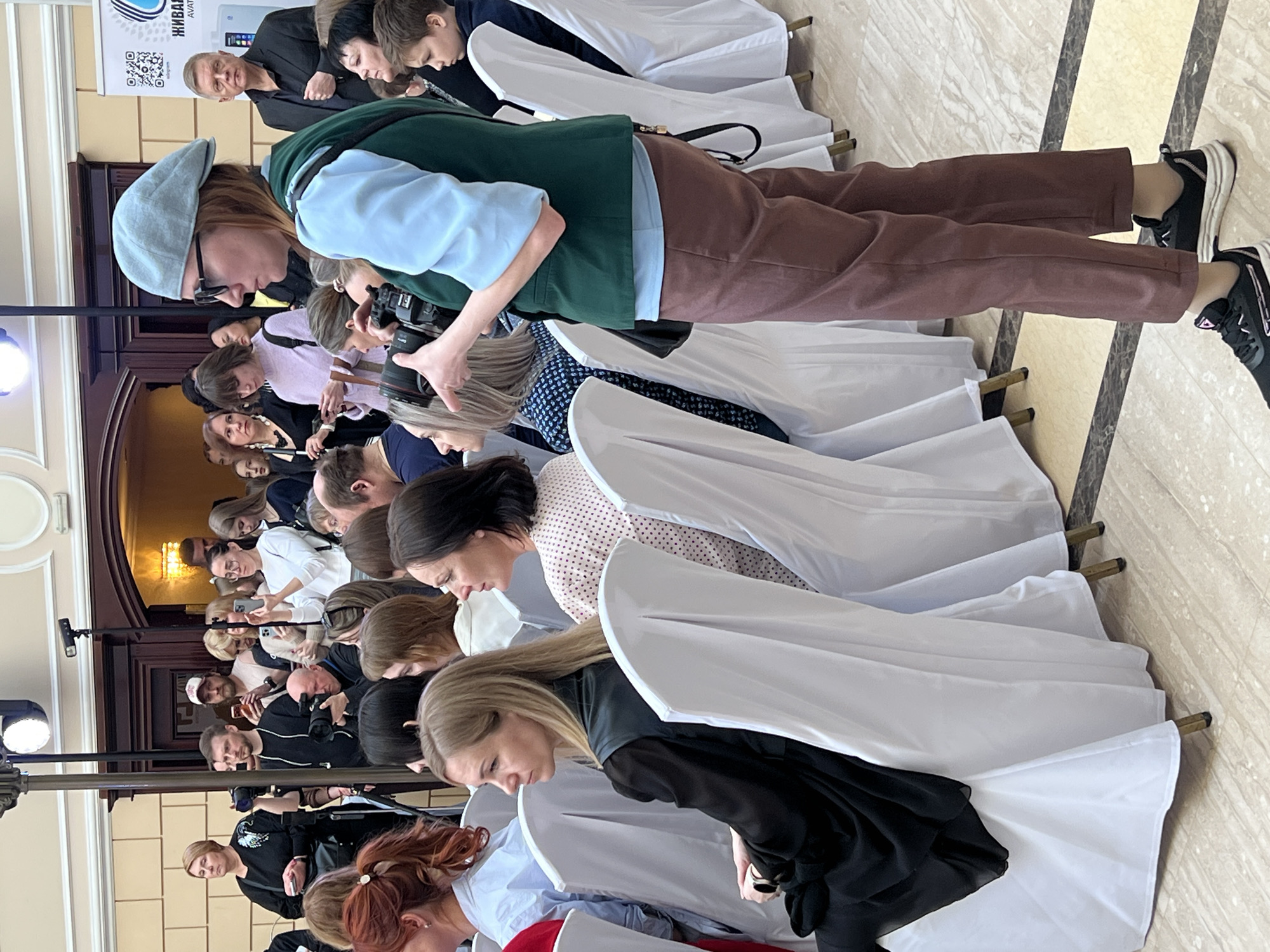 A wide shot shows a diverse audience of people seated in rows of chairs, likely attending a presentation. Most of the chairs are covered with white fabric. Individuals are looking down at their phones or devices. The person in the foreground is holding a camera. The location is in Saint Petersburg, Russia.