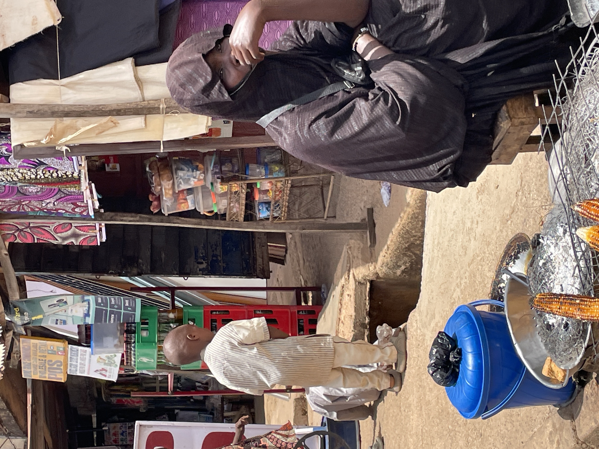 The image shows a street scene in Bukuru, Nigeria. A woman wearing a dark, patterned garment and head covering sits on a bench next to a small shop. The shop displays various goods, including packaged snacks and drinks. In front of the shop, a boy wearing a light-colored, striped shirt stands. Nearby, there's a blue bucket and a metal container holding corn on the cob. The environment appears to be outdoors with natural ground visible.
