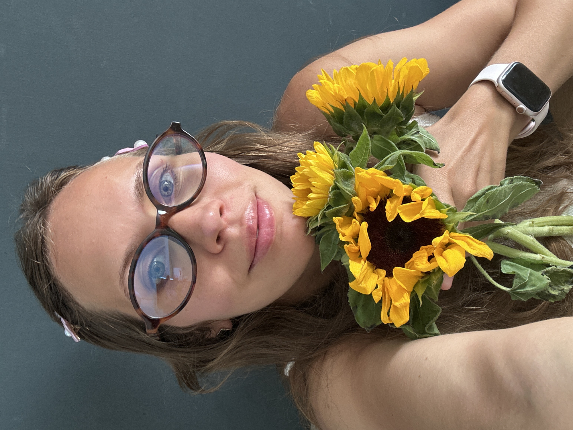 A young woman with light skin and light brown hair is holding a bouquet of sunflowers. She is wearing eyeglasses with tortoise-shell frames and a light-colored smartwatch. A hair clip with small pink and white flowers is visible in her hair. The background is a muted gray color. The scene is likely indoors due to the even lighting. There is no visible indication of the location being Fethiye, Turkey.