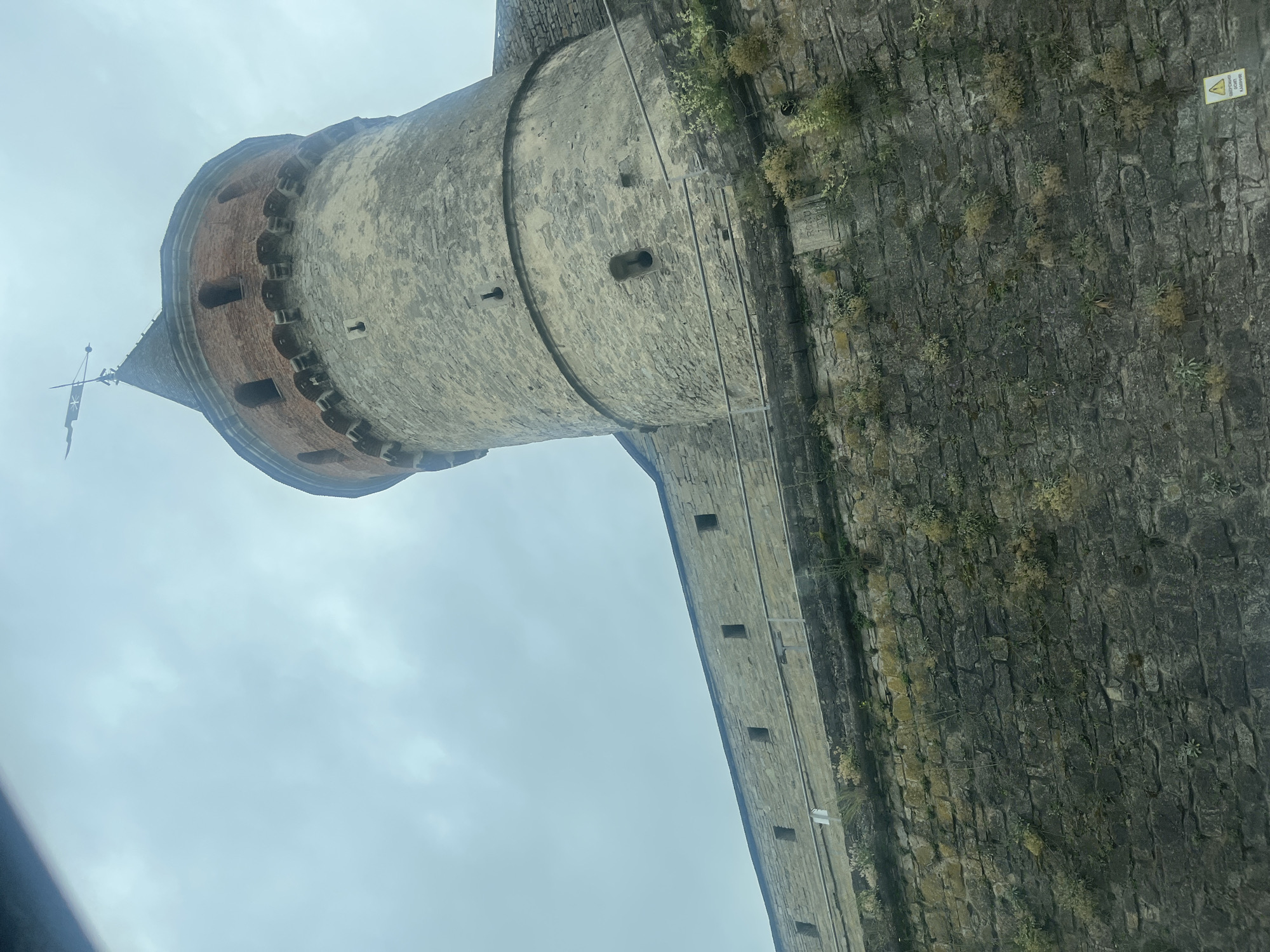 The image shows the Kamianets-Podilskyi Castle in Kamianets-Podilskyi, Ukraine. It is a view from below looking up at a large, stone tower with a conical roof and a weather vane at the top. The tower is part of a larger castle complex, with a long, stone wall extending to the left. The stone walls are weathered and show signs of age, with vegetation growing in the crevices. The sky is overcast and appears to be a pale blue or gray. There are small, rectangular openings in the stone walls, suggesting defensive features. A yellow triangular warning sign with text in Cyrillic script is visible in the lower right corner of the image.