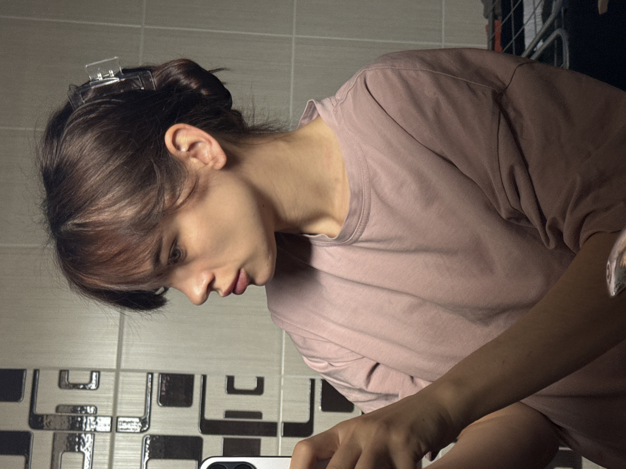 A woman with brown hair pulled back with a clear hair clip is looking down and to the left. She is wearing a light pink t-shirt. The background includes a wall with light gray tiles and dark brown geometric designs. The scene is lit with artificial light. The image contains objects like a mobile phone. The location is a bathroom. There is no verifiable evidence to determine the city of Rostov-on-Don, Russia.