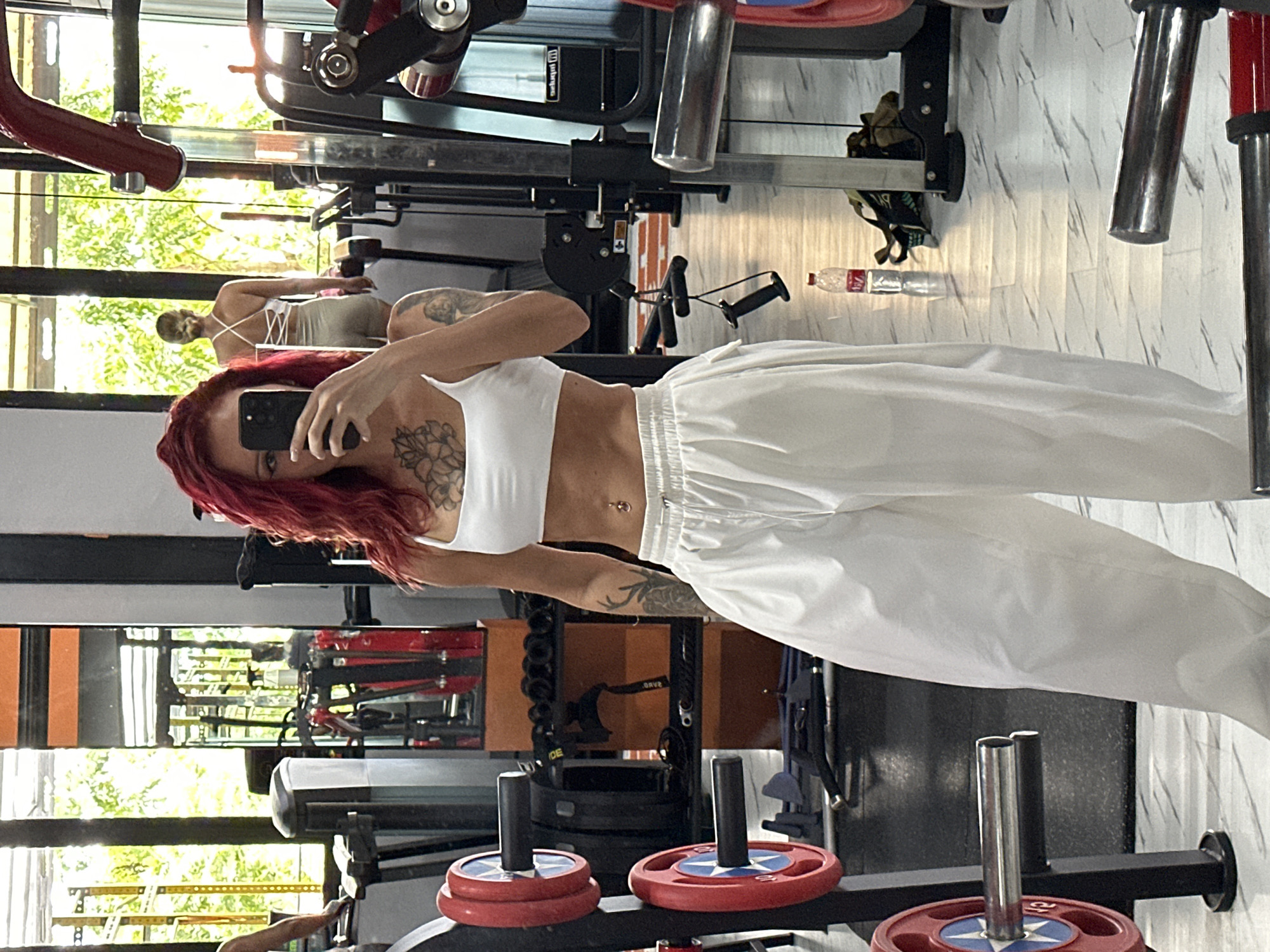 The image shows a young woman in a gym, seemingly taking a selfie in a mirror. She has red hair, fair skin, and tattoos on her arms and chest. She is wearing a white sports bra and white sweatpants. A second woman, with short blonde hair, is reflected in the mirror behind her. The gym is located in North Kuta, Indonesia. Various pieces of gym equipment are visible in the background, including weight machines, free weights, and exercise accessories. The gym has large windows, which let in natural light and offer a view of the outdoors. The flooring is a white marble-like tile. A water bottle is on the ground in the background.