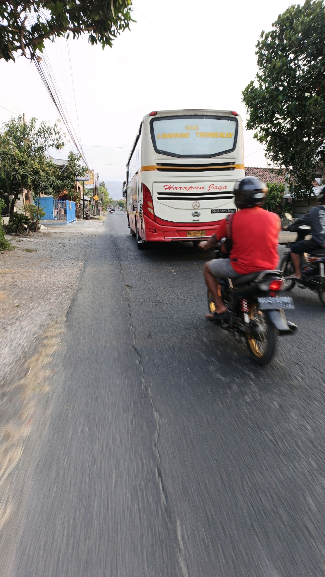 A low-angle, eye-level, full shot captures a street scene in Pojok, Indonesia, with a large white and red bus dominating the background. The bus, with "PATAS LANGSUNG TRENGGALEK" written on its upper windshield and "Harapan Jaya" on its side, is positioned on the right side of the road, moving away from the viewer. In the foreground and mid-ground, two motorcycles are in motion. The first motorcycle, directly in front of the bus and closest to the viewer, is ridden by a person wearing a red shirt and gray shorts, with a black helmet. The second motorcycle is further back and to the right, with its rider mostly obscured. The asphalt road is cracked and worn, with a shoulder of gravel and vegetation on the left. Trees with dense foliage line the left side of the road, and buildings and houses are visible in the distance on both sides. The sky is overcast, suggesting a daytime scene with diffused light. The motion blur indicates that the photograph was taken from a moving vehicle.
